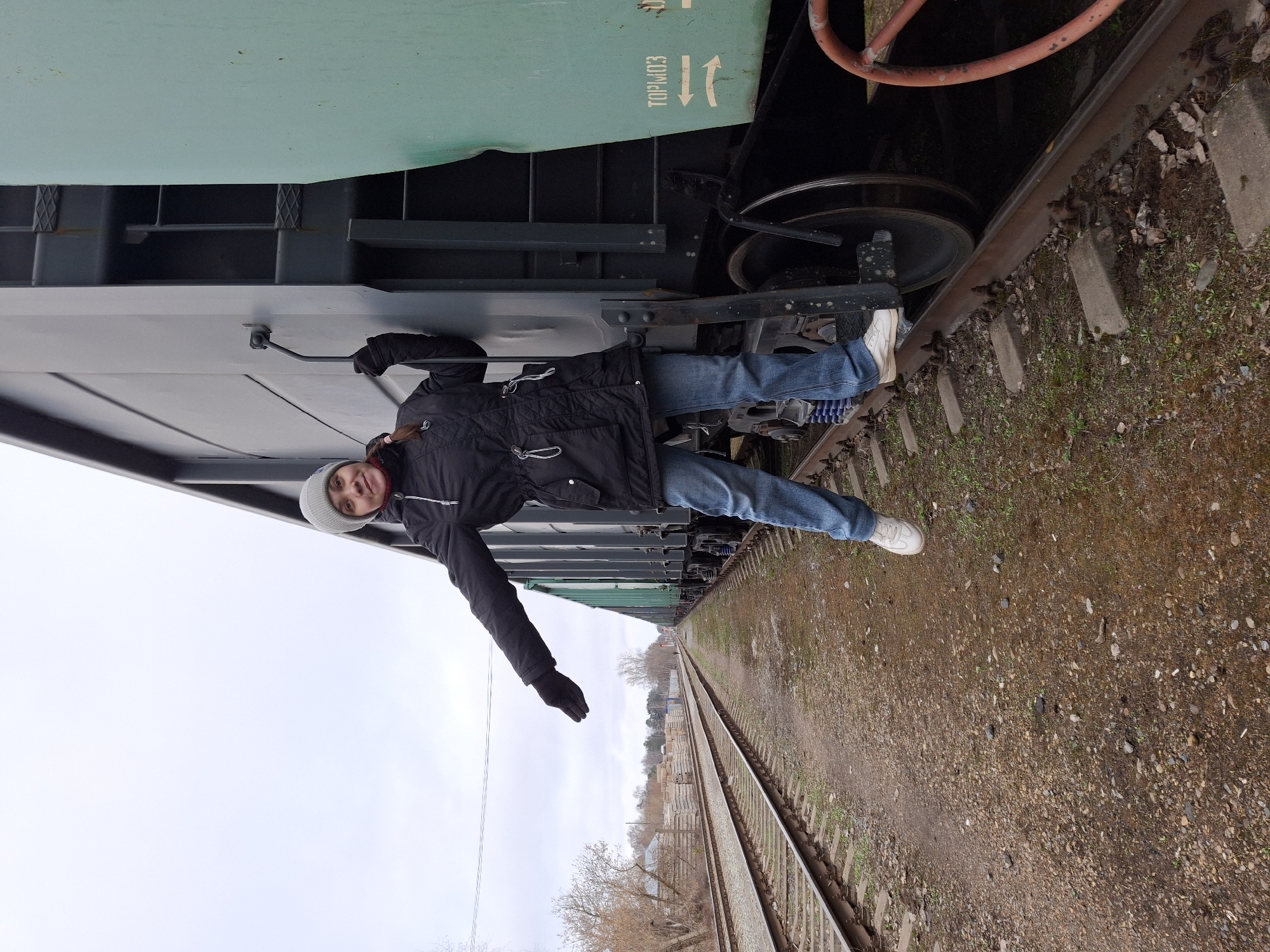 The image displays a person lying horizontally against the side of a large train car, with train tracks and an overcast sky in the background. The photograph is rotated 90 degrees clockwise, giving the illusion that the person is suspended sideways along the length of the train car.
The main subject is a person wearing a black quilted jacket, blue jeans, white sneakers, black gloves, and a light grey beanie. They are positioned with their head towards the top-left of the frame and their body stretched out, their left arm extended downwards. A slight smile is visible on their face, suggesting a playful or posed activity.
The train car, which takes up the left side of the frame, has a green upper section and a darker, possibly black or dark grey, lower body and undercarriage. Visible beneath the car are train wheels and various structural components. On the green section of the train car, white text reads "TOP.W.0.3" followed by two white downward-pointing arrows. A rusty, reddish-orange metal bar is also visible near the top-right corner of the train car.
To the right of the train car, two parallel sets of railway tracks extend into the distance. The ground between and around the tracks is covered in gravel ballast with patches of green vegetation. The sky is uniformly light grey, indicating an overcast day with diffused lighting and no strong shadows. In the distant background, a line of bare trees and some low-lying structures can be seen, suggesting an outdoor, possibly semi-rural or industrial setting during a cooler season.