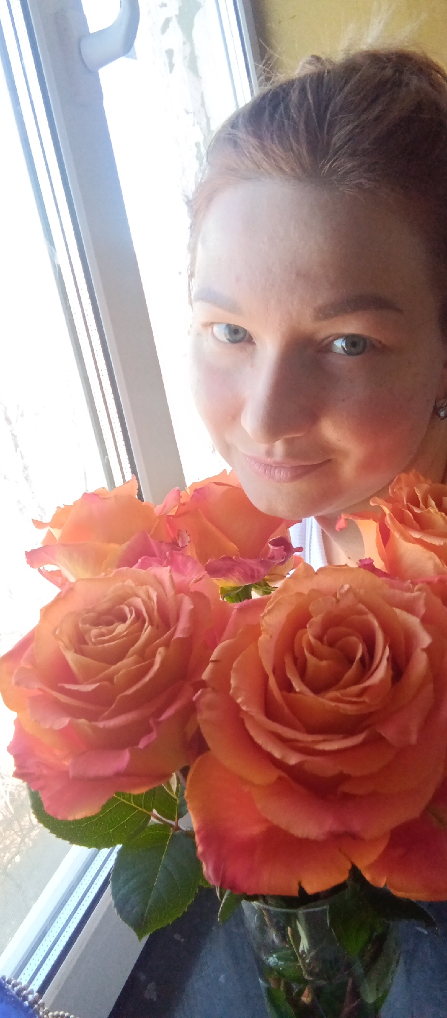 The image is a selfie of a woman holding a bouquet of orange roses, presumably inside a building in an unidentified city. The woman has fair skin, red hair pulled back, light-colored eyes, and is wearing earrings. She is smiling slightly. The roses are a vibrant orange color with hints of pink on the edges of the petals, and green leaves are visible. The background features a window with white frames and a blurred view outside. The specific location within the unknown city is indiscernible.