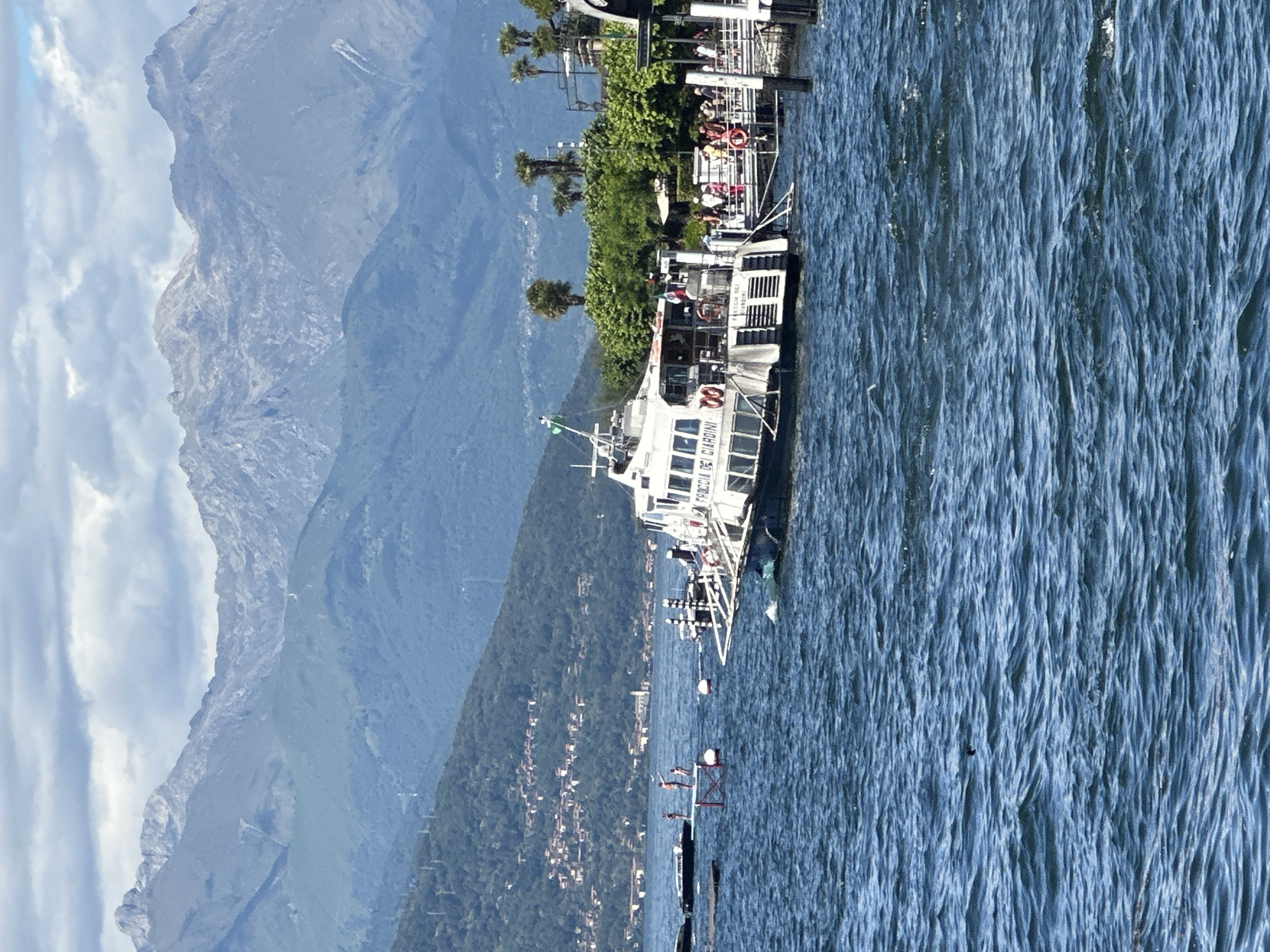 The image depicts a ferry docked at a pier on a body of water, with a mountainous landscape in the background. The ferry, named "FRECCIA DEI GIARDINI," is white with multiple windows and appears to be carrying passengers. Several people are visible on the pier, some boarding or disembarking the ferry. The water is choppy, with small waves. In the distance, a hillside town with red-tiled roofs is visible against a backdrop of lush green trees and imposing mountains under a cloudy sky. The scene is set in San Giovanni, Italy, likely on a lake given the mountainous terrain and ferry operation.