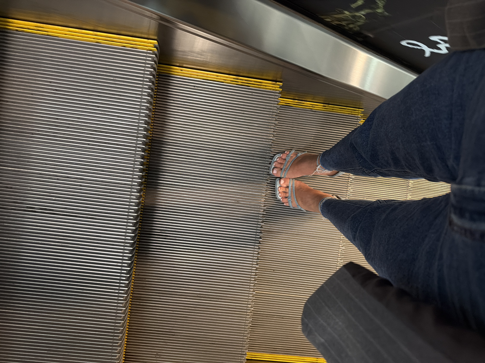 A person stands on an escalator in Vnukovo, Russia. The escalator steps are made of metal with yellow safety strips along the edges. The person is wearing blue jeans and light blue jeweled sandals. A dark-colored bag is held in the person's left hand. The background shows the metal and yellow components of the escalator.