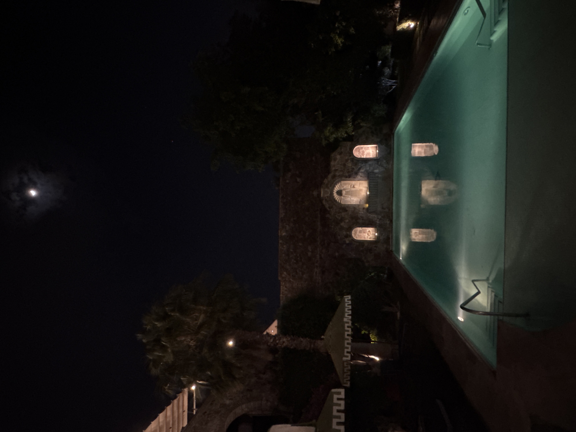 Here is a description of the image:
The image shows an illuminated swimming pool at night. Above the pool is a structure with lit alcoves. To the left of the building is a dark sky, lit by the moon. Palm trees are also visible to the left, and the building has a crenellated wall on the left. The image orientation is such that the sky and moon are on the left side of the frame, and the pool is on the right. It is at night in the city of San Miguel de Allende, Mexico.