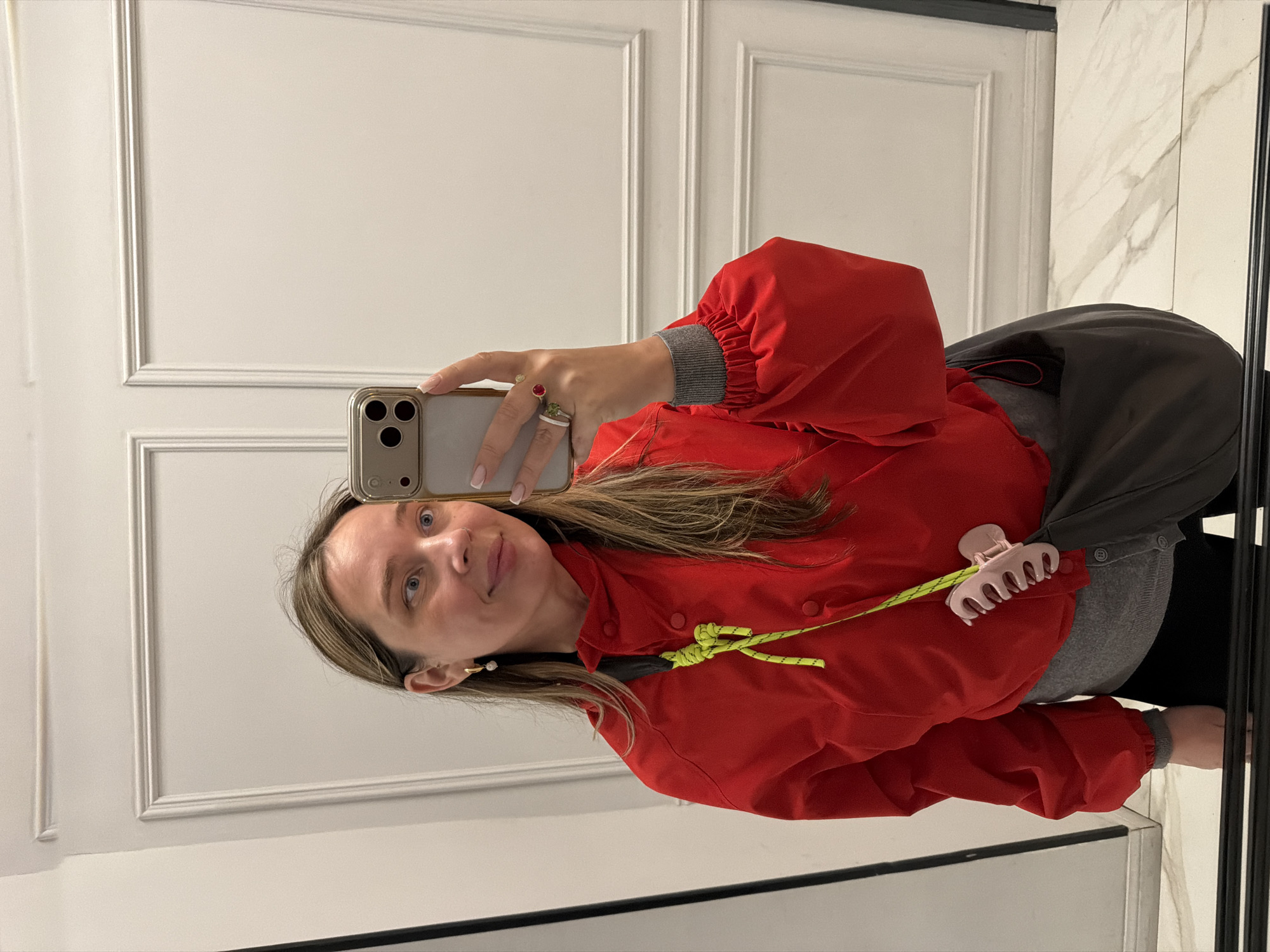 A woman is taking a selfie in what appears to be an interior space, possibly a bathroom or entryway, based on the presence of a mirror, white paneled walls, and a marble-like surface. She is holding a smartphone in her right hand and facing the mirror at an angle, and is wearing a red jacket with neon yellow drawstrings, a gray undershirt, and rings on her fingers. She has light-colored hair and a small pearl earring in her left ear. A light-pink hair clip is visible on the left side of her jacket. A black bag is slung over her shoulder. The location in Moscow, Russia cannot be determined from the image alone.