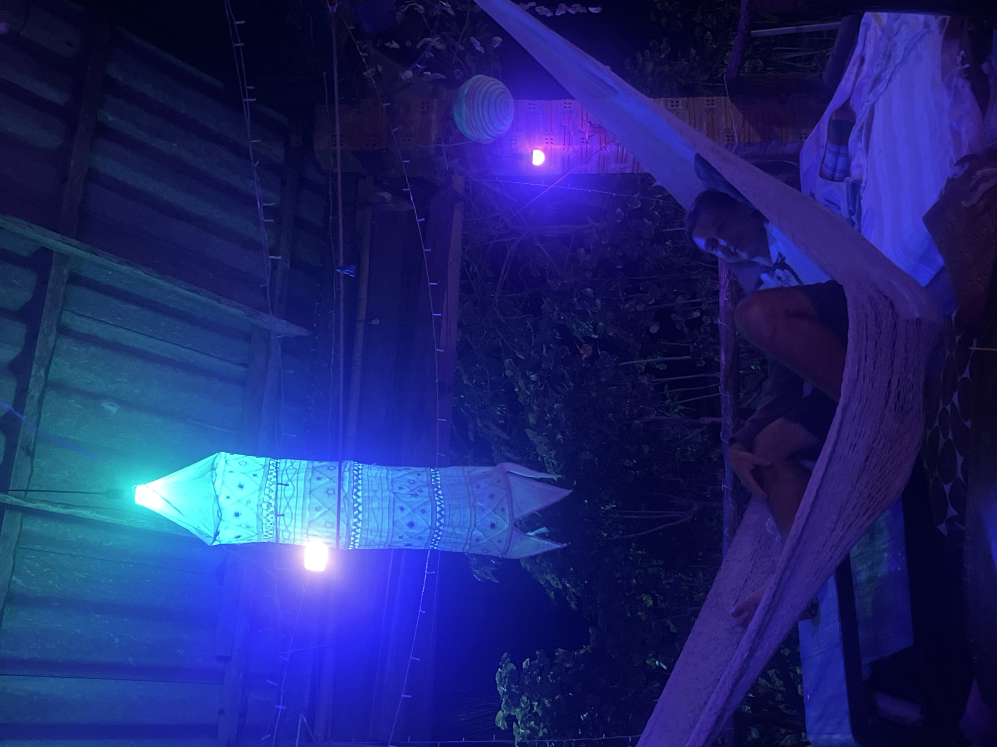 The image captures a dimly lit, outdoor scene at night. The primary subjects are a person relaxing in a hammock and several decorative lanterns. The person is a young individual, appearing to be male, dressed in dark shorts and a light-colored t-shirt, and is reclined in a woven hammock. The hammock is suspended outdoors, likely in a tropical or humid environment, given the lush dark foliage visible in the background.
Dominating the left side of the image is a long, cylindrical lantern emitting a bright green-blue light. This lantern is decorated with intricate patterns, suggesting it's made of fabric or paper. Further in the background, another spherical lantern with striped patterns is visible, illuminated by a soft pinkish-orange light, and a smaller, diffused light source that appears to be purple. The overall lighting is moody and atmospheric, with a strong presence of blue and green hues from the lanterns.
The setting appears to be a rustic, possibly outdoor living space or recreational area. Wooden structures with corrugated roofing are visible on the left, suggesting a shelter or building. Hanging wires and strings indicate the presence of multiple lights and decorations. The ambiance is relaxed and possibly festive, with the person in the hammock appearing at ease. The time of day is clearly night, and there are no indications of weather conditions. No visible text is present in the image.
