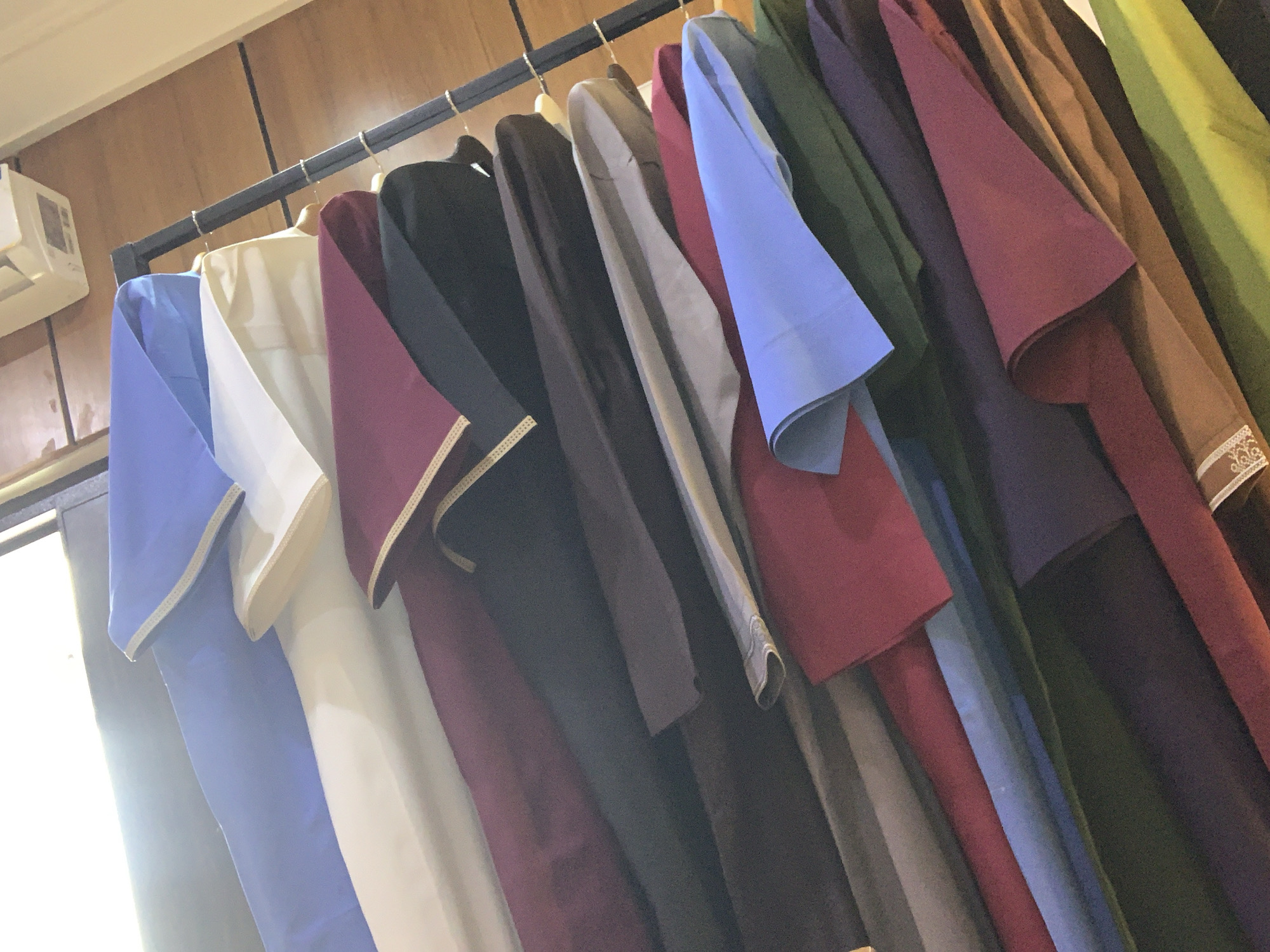 The image shows a rack of clothing hanging in what appears to be a retail setting. The clothing items are of various colors, including blue, white, maroon, black, green, and yellow. They are all similar in style, suggesting they are part of a collection or uniform. The background is a wooden paneled wall. On the left side of the image, a white air conditioning unit is visible mounted on the wall. The scene is well-lit, likely from natural light coming from a window or door on the left, which is partially visible and out of focus. The overall impression is of a shop or storage area for garments.