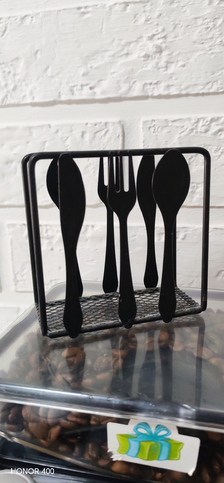 A cutlery holder containing several pieces of black flatware (spoons, forks, and knives) sits on top of a container filled with coffee beans. The container has a sticker with a cartoon drawing of a present. The background is a white brick wall. The text "HONOR 400" is visible in the lower left corner of the image. The specific location in the city of Noviy, Russia, cannot be determined from the image.