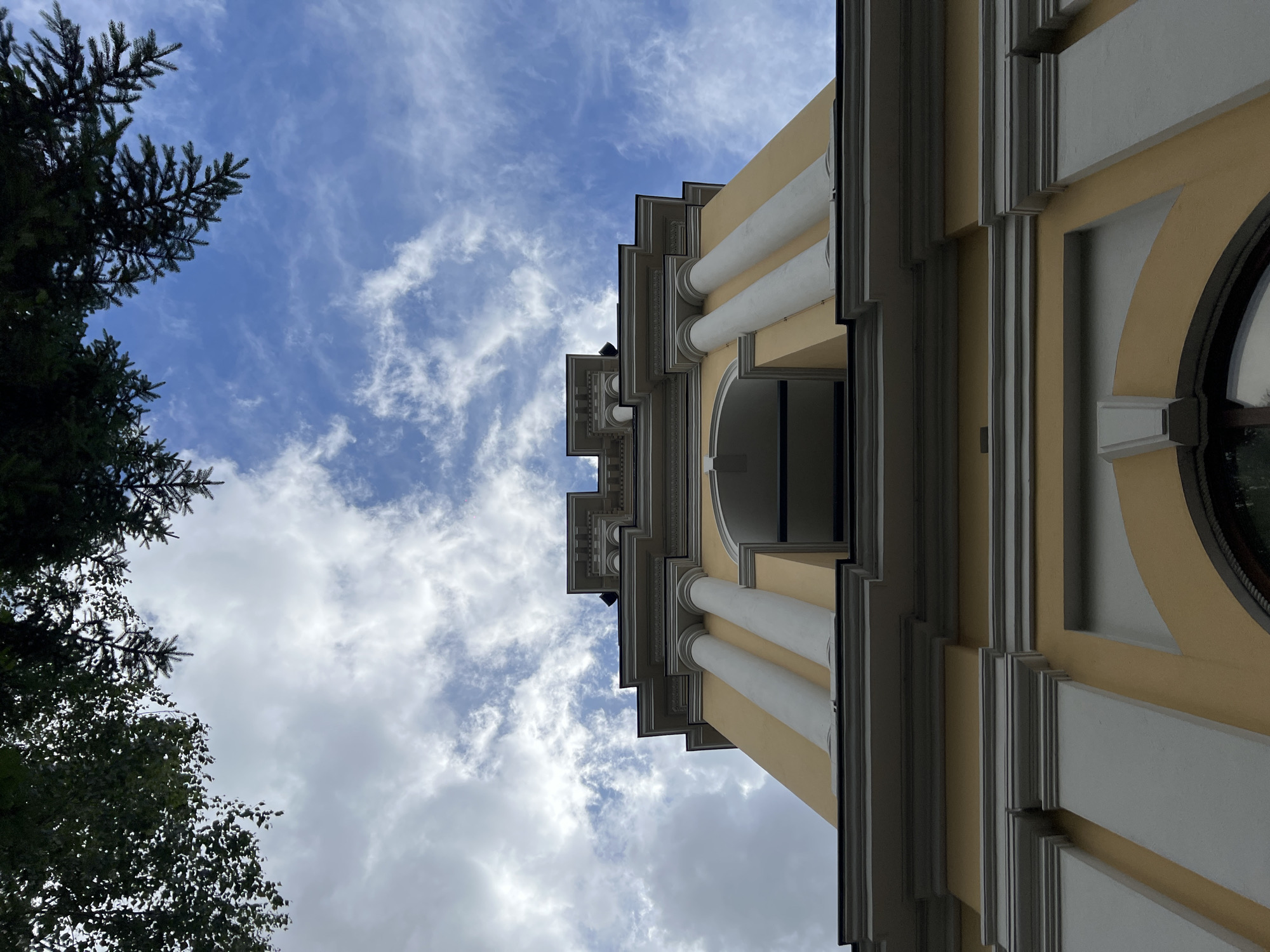 The image shows a low-angle view of a yellow and white building with classical architectural features, including columns and decorative moldings, set against a blue sky with scattered clouds. Tree branches are visible on the left side of the frame, partially obscuring the sky. The building's structure includes an open archway supported by columns, suggesting it could be part of a bell tower or similar structure. No people are visible in the image.
Although a specific building cannot be identified from this image, the architecture suggests it may be in the city of Moscow, Russia.