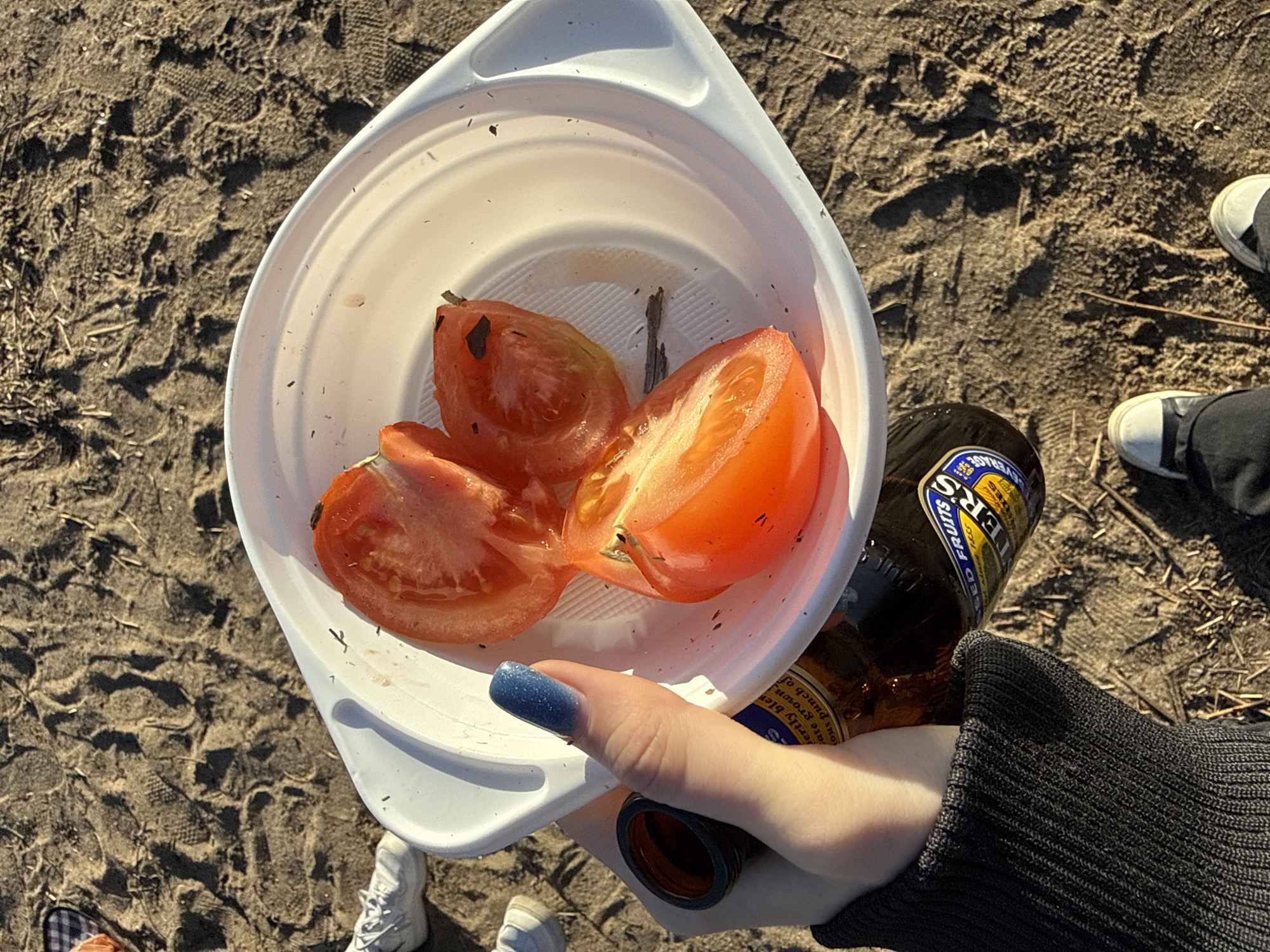The image shows a first-person view of a person holding a white plastic dish with three tomato slices and a bottle of beer. The person's hand, adorned with blue glitter nail polish, grips the beer bottle's neck. The person is wearing a dark gray sweater. The background consists of a sandy surface with scattered debris and footprints, along with glimpses of shoes and clothing suggesting other people are nearby. The location is likely near Toksovo, Russia.