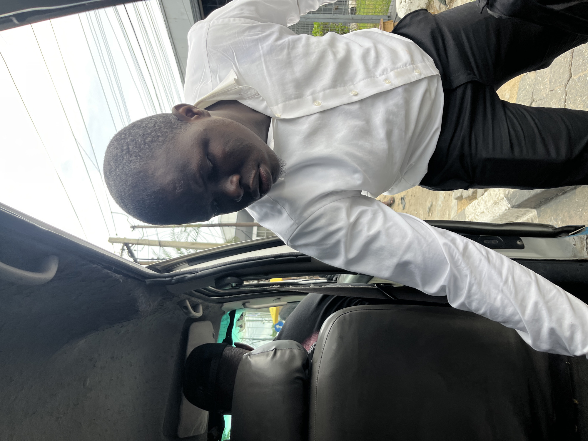 A man is leaning into the open roof of a car. The man has dark skin, short dark hair, and is wearing a white long-sleeved button-down shirt and black pants. He appears to be reaching down inside the vehicle. The interior of the car is visible, including seats and other interior elements. Overhead power lines are visible in the background. The location is Itirin, Nigeria.