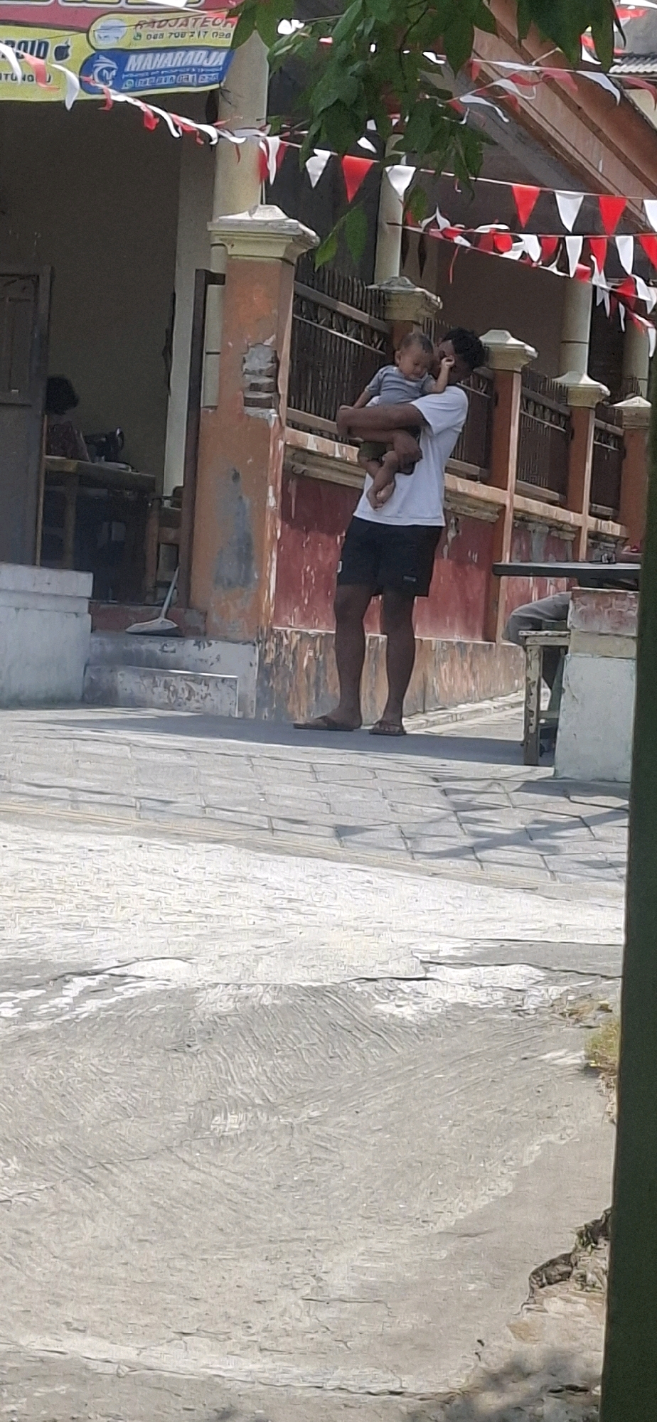 In Blitar, Indonesia, a man in a white t-shirt and black shorts is holding a baby. The baby, wearing a gray onesie, is being cradled in the man's arms. The man is standing on a paved walkway in front of a building with a red and orange facade. The building has a fence in front of it, and colorful triangular flags are strung across the scene. In the background, a person is visible inside the building, seated at a table with what appears to be a sewing machine. A broom leans against the wall near the entrance. The ground is paved with stone tiles.