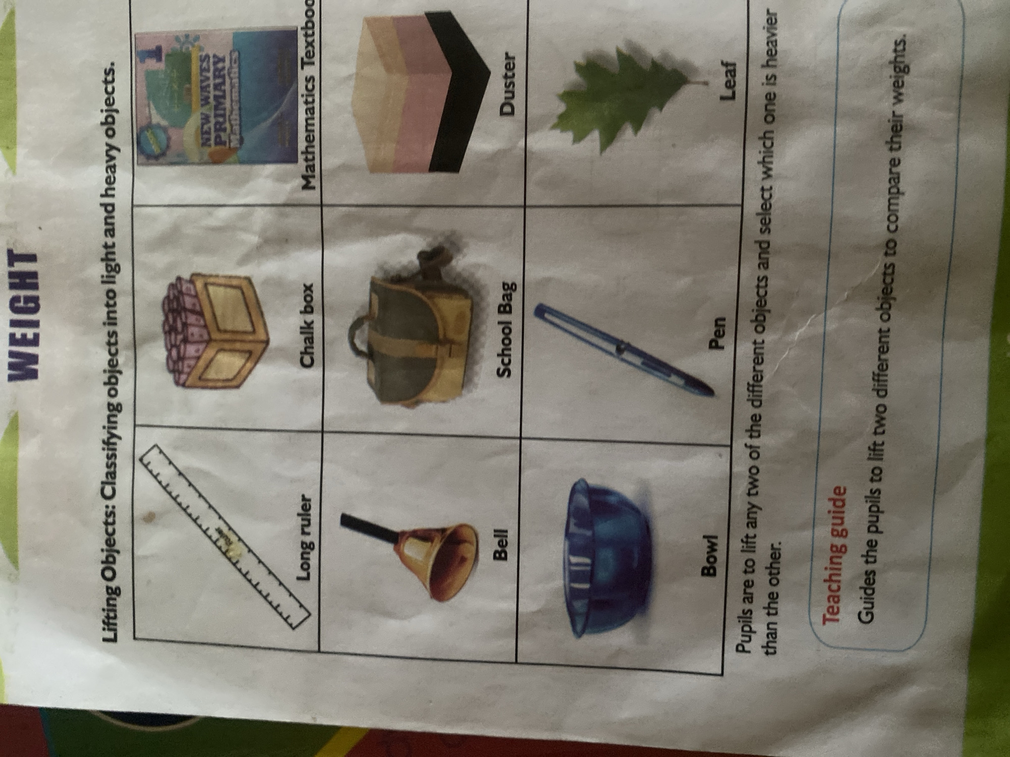 The image is a page from a children's educational worksheet, likely for a primary school class, focused on the concept of "Weight" and classifying objects as light or heavy. The page is divided into a grid of six cells, each containing an illustration of an object and its label. The objects depicted are: a long ruler, a chalk box, a mathematics textbook, a bell, a school bag, and a duster. Below this grid, there are three more cells: a bowl, a pen, and a leaf. Underneath these are instructions for pupils to lift two different objects and select the heavier one. Finally, there is a "Teaching guide" section with further instructions for the teacher. The worksheet has a slightly wrinkled appearance, suggesting it's a printed page that has been handled. There is no indication of the city of Jalingo, Nigeria, or any specific location within it.