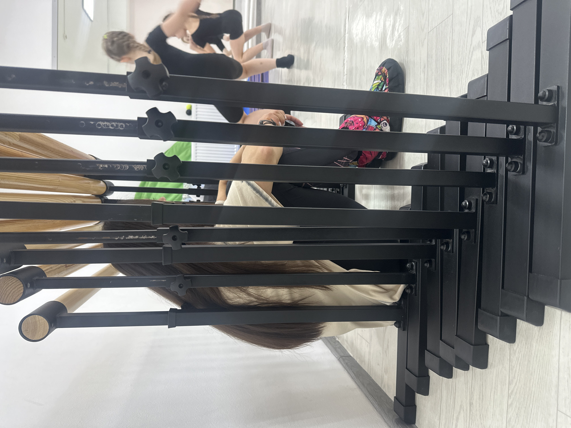 This image depicts an indoor scene within what appears to be a dance or fitness studio in Votkinsk, Russia. The foreground is partially obscured by a stack of black metal chairs or stands, arranged vertically. To the left, several wooden ballet barres are mounted on black metal supports with adjustable knobs.
Behind these structures, multiple individuals are present. In the midground, a person with long brown hair is seated on the light-colored floor, leaning against the stacked chairs. They are wearing a light-colored top, and one foot, adorned with a brightly patterned sneaker, is visible. Another person's arm and hand, holding what appears to be a mobile phone or small device, can be seen between the black bars.
In the blurred background, two figures are engaged in activity. Both are dressed in black athletic wear. One individual, with light-colored hair, is seen bending forward. The other figure is also in a dynamic pose, possibly stretching or performing a dance movement, with their legs extended. One leg has a white sock, the other a black sock. The setting features light-colored flooring and plain white walls, suggesting a clean, functional studio space. A window or light source in the upper left background indicates a well-lit interior. The overall impression is of a practice session or class. No discernible text is visible in the image.