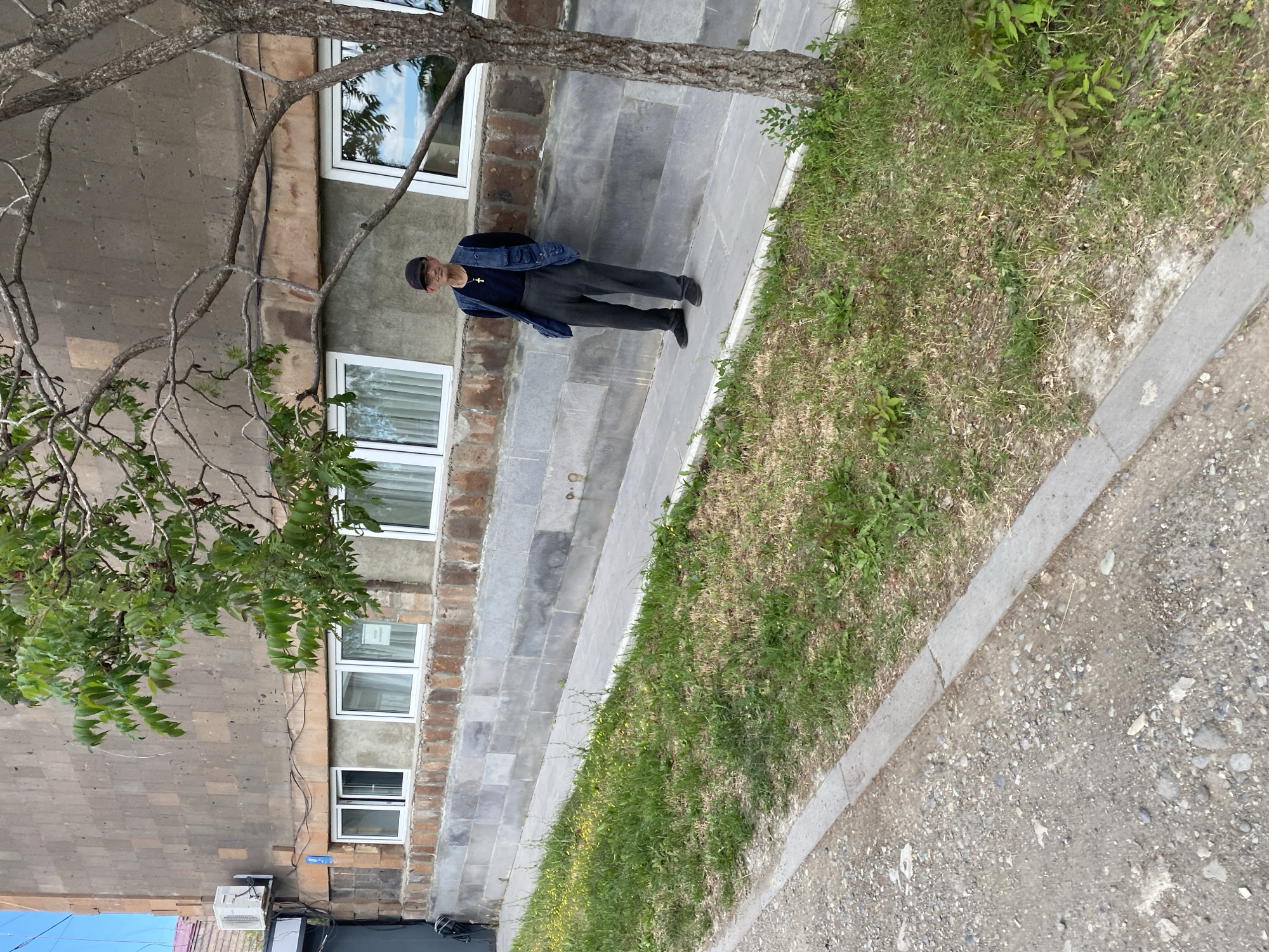 An elderly man stands on a paved walkway in front of a multi-story building in Gyumri, Armenia. The building has a rough, brown-toned stone facade with a row of white-framed windows running horizontally across its midsection. The walkway is bordered by a strip of green grass with scattered yellow wildflowers, which then transitions to a gravel-covered area. A tree with sparse green leaves and a rough bark trunk stands to the right of the man, partially obscuring the building. The man is wearing a dark baseball cap, a dark long-sleeved shirt, a dark blue vest, and dark grey pants. He is wearing black shoes and a cross pendant around his neck. He stands with his hands behind his back, looking towards the camera.