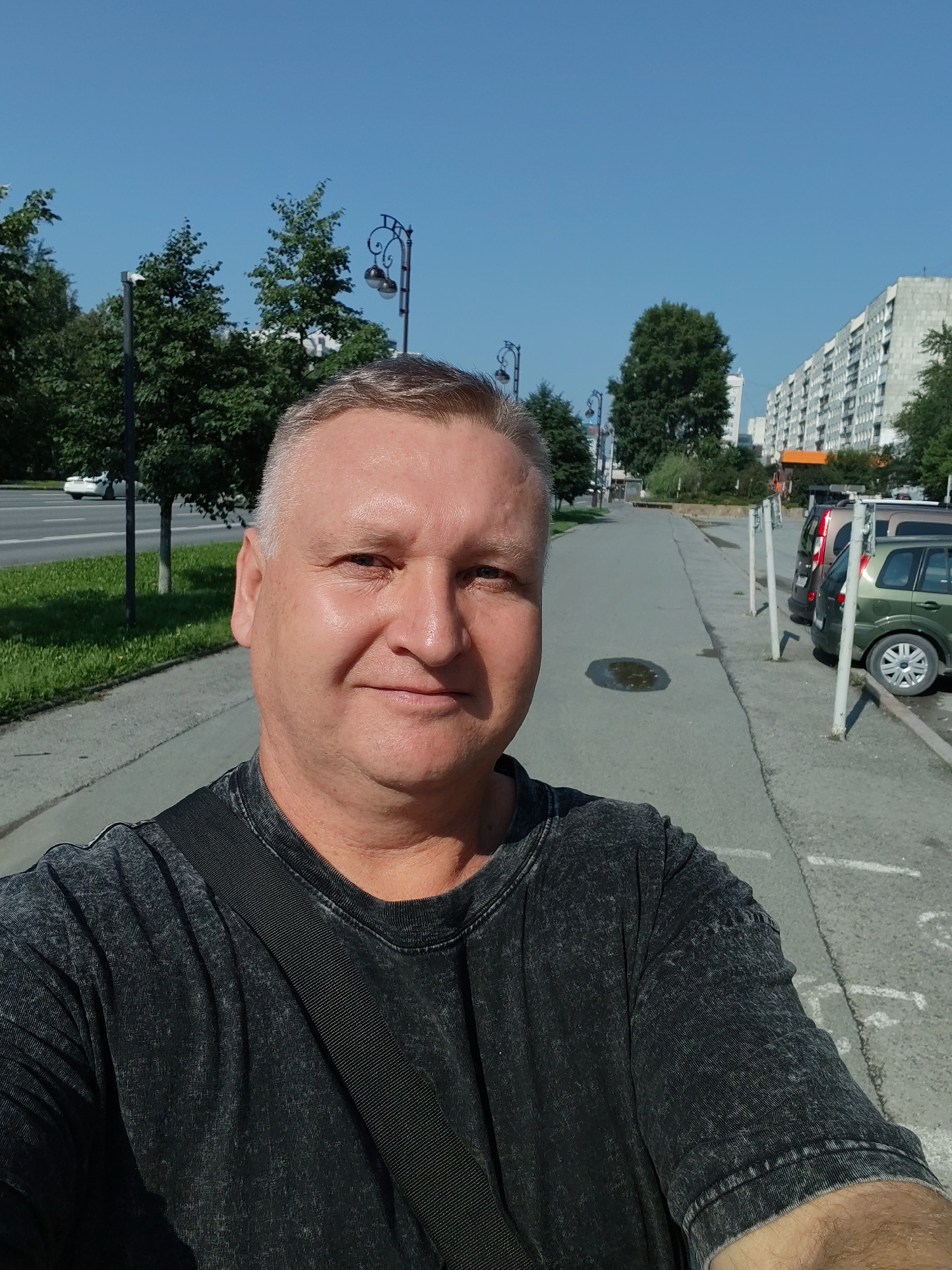 A medium shot, selfie-style photograph shows a Caucasian man with short, graying hair and fair skin. He is wearing a dark gray, t-shirt with a black strap across his chest. He is standing on a sidewalk in front of a street, with a row of parked cars to his right. In the background, there are trees, streetlights, and a tall apartment building. The sky is clear and blue. The man is looking directly at the camera and smiling. The overall lighting suggests a sunny day.