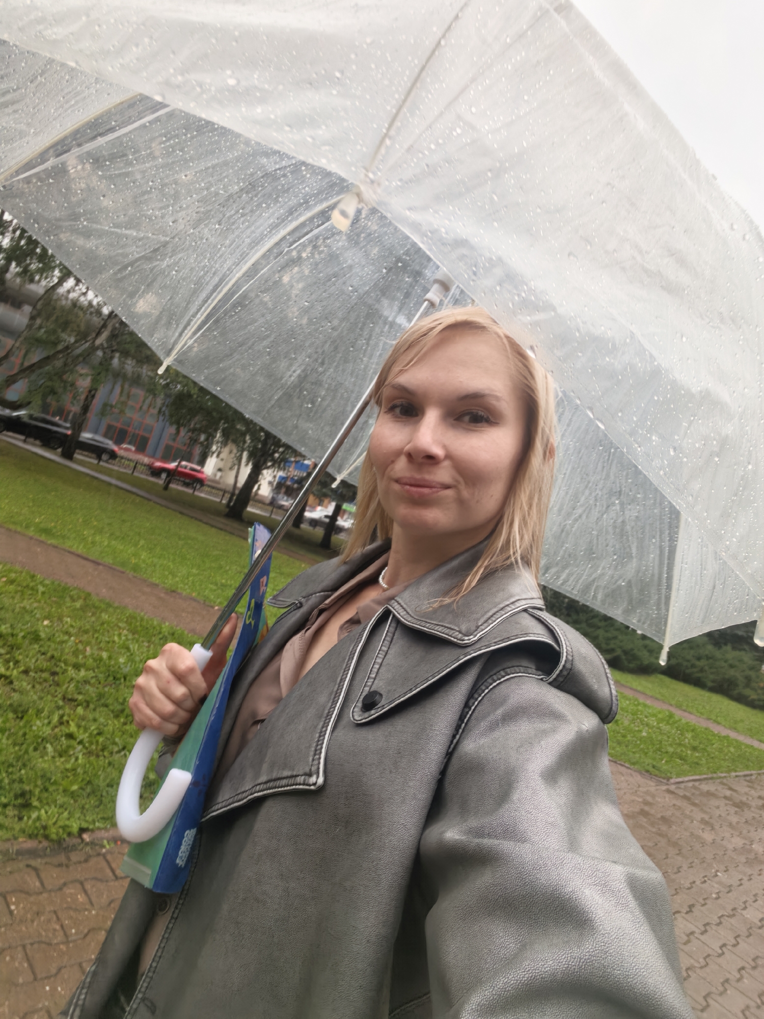 A woman with blonde hair is holding a clear umbrella with raindrops on it. She is wearing a gray, faux leather jacket and a light brown shirt. She is also holding a folder or binder with a blue and green cover. The woman is standing on a sidewalk made of interlocking pavers, and there is a grassy area to her left with trees and buildings in the background. Cars are visible on the street. The scene appears to be in a city environment, possibly in Sterlitamak, Russia, given the contextual prompt. The weather is overcast and wet, indicating it is raining.