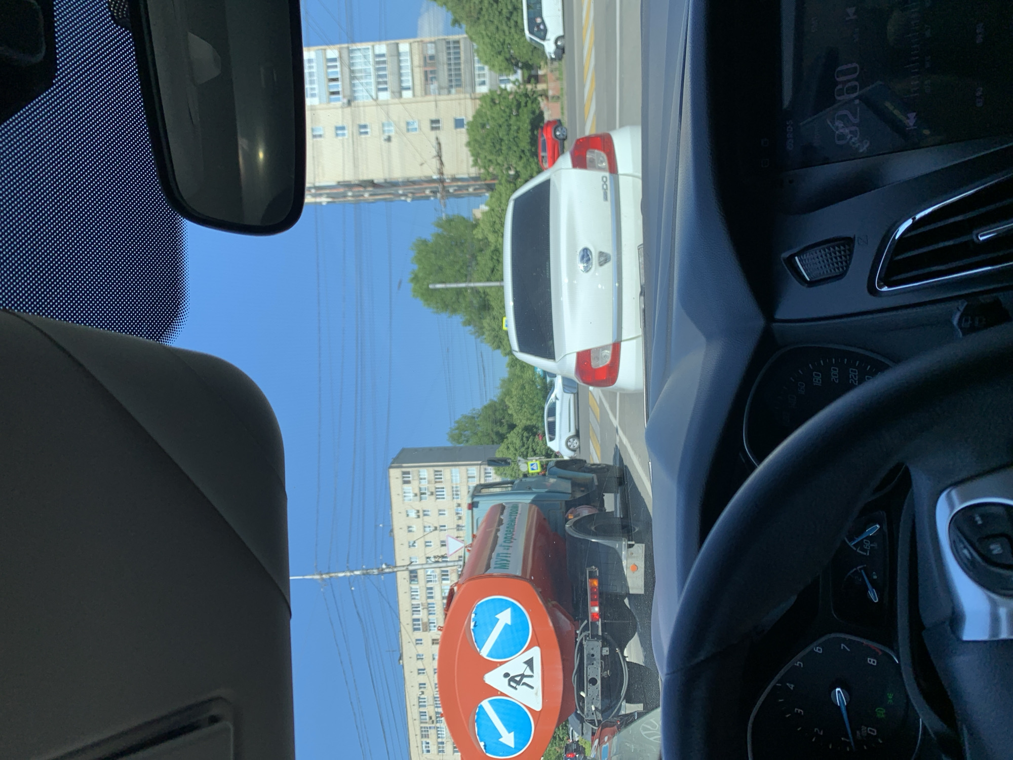 In Stavropol, Russia, on a sunny day, the image is captured from inside a car. The view is through the windshield, showing traffic on a city street. A large orange tanker truck with blue directional arrows and a worker symbol on its rear is prominent. The truck displays Cyrillic text, possibly indicating the company name. Behind the truck, a white sedan is visible, followed by a red car and a white van. In the background, multi-story apartment buildings and trees line the street. The car's dashboard and steering wheel are partially visible in the foreground. The sky is clear and blue.
