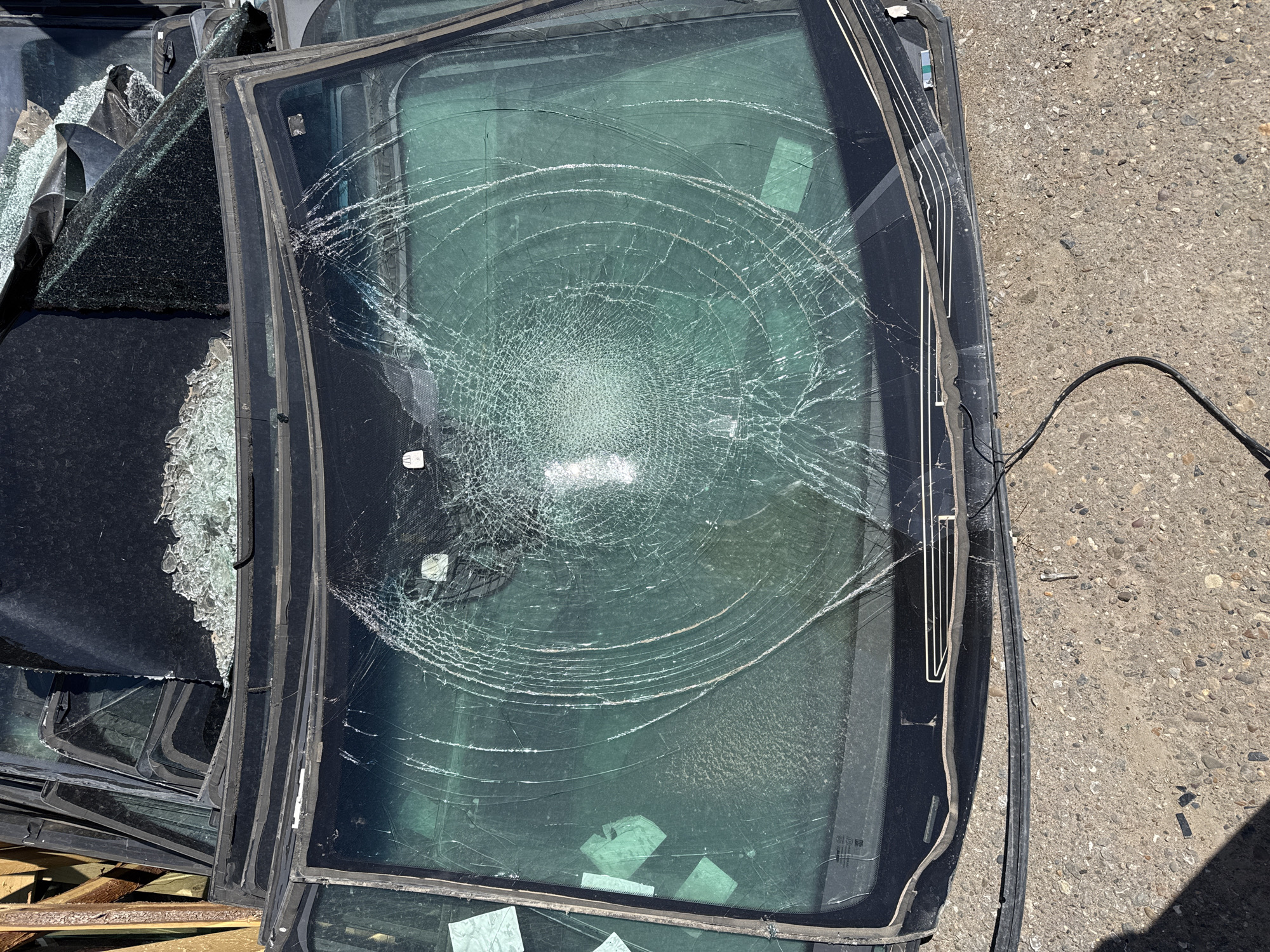 A stack of discarded vehicle windows is piled on a wooden pallet atop a gravel surface, presumably outdoors. Many windows are broken and spider-web cracked. A black cable lies adjacent to the glass pile. The location is not discernable as Naberezhnye Chelny based solely on the visual content of the image.