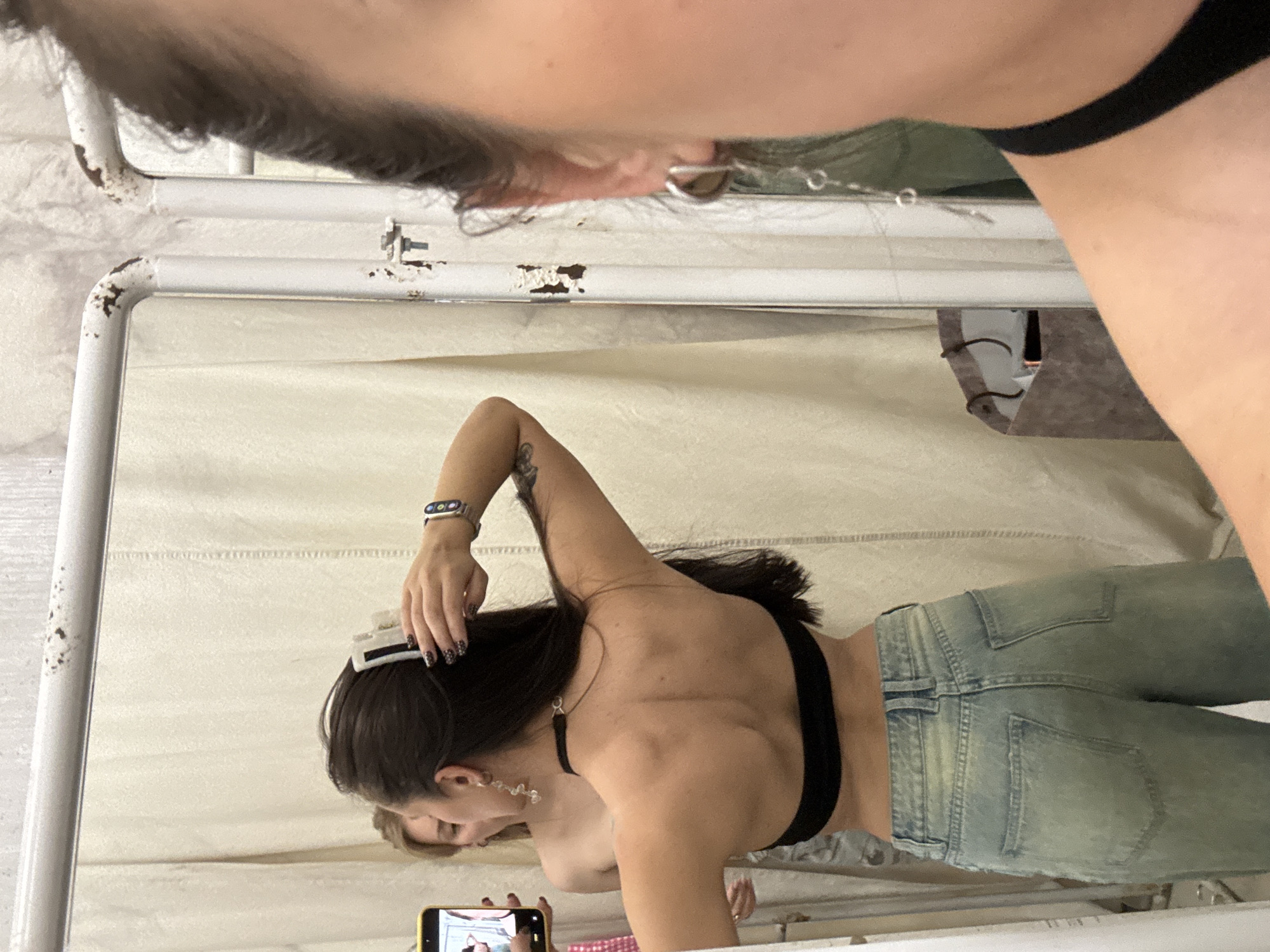 An image, rotated 90 degrees clockwise, captures a mirror selfie being taken indoors, likely in Moscow, Russia. The main subject is a person reflected in a large, rectangular mirror with a white, somewhat rusted metal frame. This person has dark hair, wears a black bra or crop top, and light blue jeans. A watch is visible on their left wrist, and they wear dangling, clear earrings. Their right hand holds a yellow-cased smartphone, pointed towards the mirror, actively taking the selfie. Their left hand is near their head, possibly adjusting hair or a white hair clip, and their fingernails display a dark, speckled pattern. The reflection shows the person’s back and a side profile.
Above the mirror, partially visible, is the head of another person (or the individual holding the camera) with dark hair and part of their upper body clad in a black garment. This individual is presumably taking the photograph of the mirror's reflection. The background behind the mirror consists of a light-colored fabric or wall. A gray or brown shopping bag with dark handles is partially visible on the right, outside the mirror's frame. The setting appears casual, with indoor lighting. No discernible text is visible.