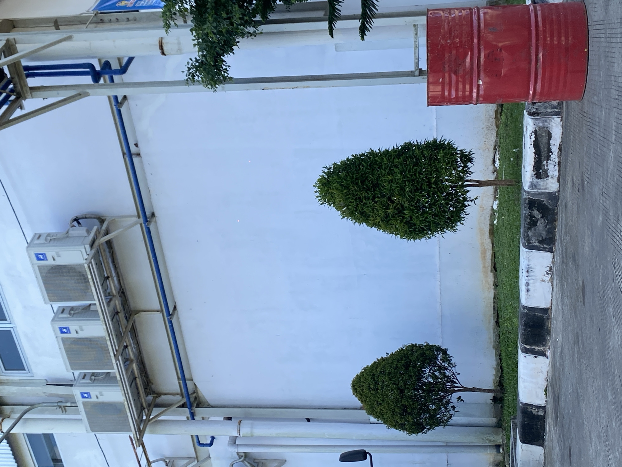In Gambut Barat, Indonesia, a red metal barrel stands to the right of the frame, positioned in front of a white building. Three outdoor air conditioning units are mounted on a metal rack attached to the building's exterior wall. Two small, cone-shaped green trees are planted in front of the building, with a grassy area behind a curb separating them from the paved ground. Blue pipes are visible running along the building's exterior. The scene appears to be an outdoor industrial or commercial area.