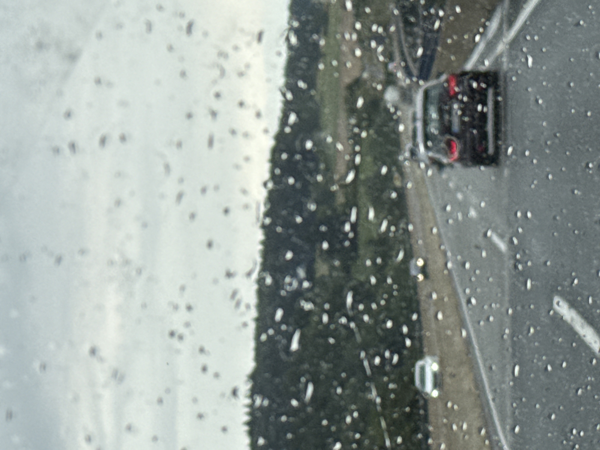 The image shows a view through a rain-streaked car windshield on a highway. In the foreground, water droplets are visible on the glass. The road ahead is gray asphalt with white lane markings. Several cars are on the highway. A dark-colored car, possibly a hatchback, is in the right lane, slightly ahead and to the right. Further ahead and to the left, a lighter-colored car is visible in the left lane. In the distance, rolling hills covered in dense green trees form the backdrop. The sky is overcast and grayish. The scene appears to be from a highway in or near Bad Lauterberg im Harz, Germany, given the presence of a highway and the type of vegetation. The weather is inclement, with rain affecting visibility.