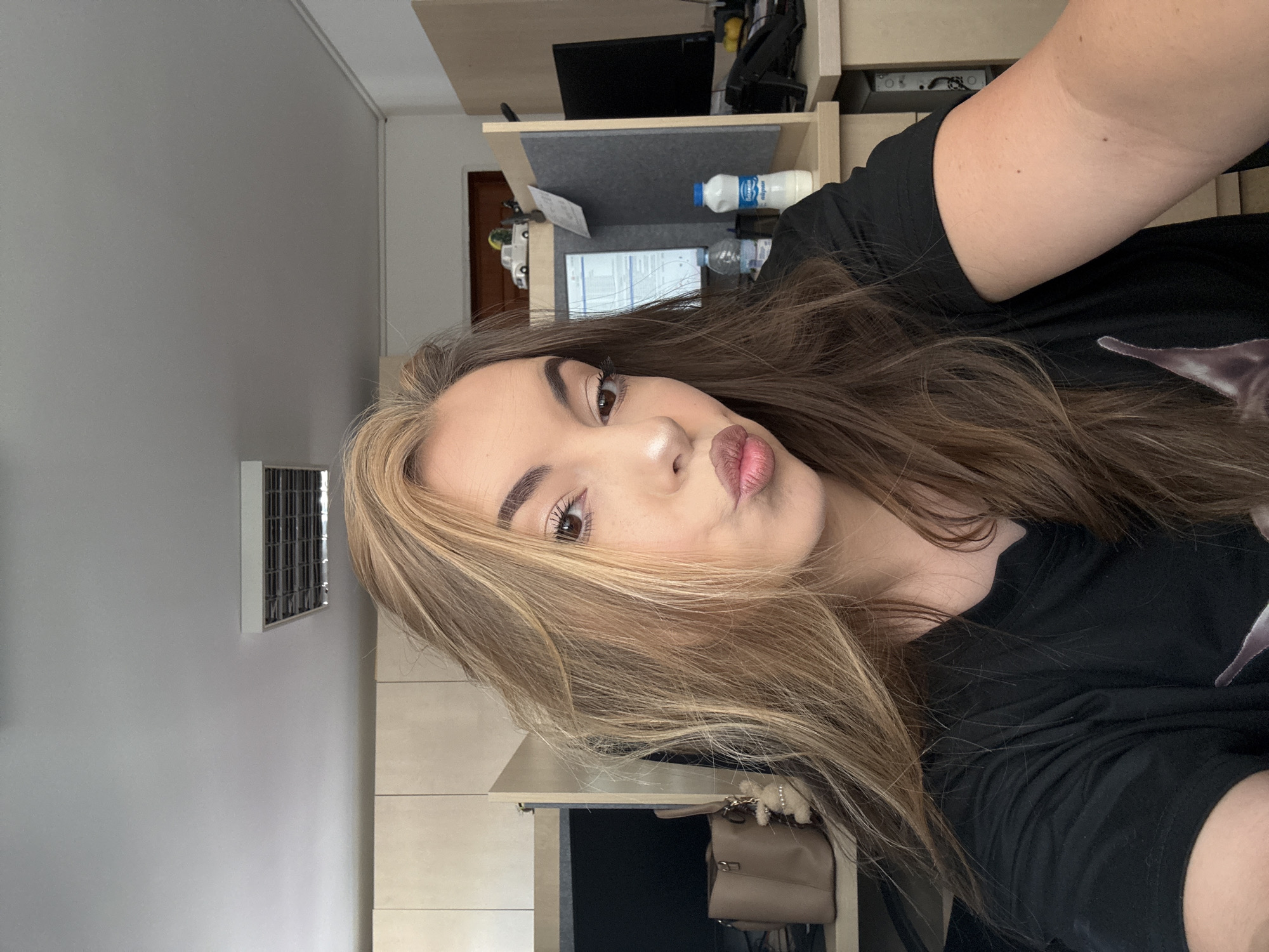 A selfie, rotated counterclockwise, shows a fair-skinned person with long, light brown and blonde hair, full eyebrows, and brown eyes. The person is wearing a black t-shirt and pursing their lips towards the camera. In the background is a light gray wall with a rectangular vent. A beige cabinet is also visible. Behind the person, there is a desk with a computer monitor, keyboard, bottle of water, and a bottle of milk. A purse is hanging on a hook. It is impossible to confirm this is the city of Sofia, Bulgaria from the image provided.