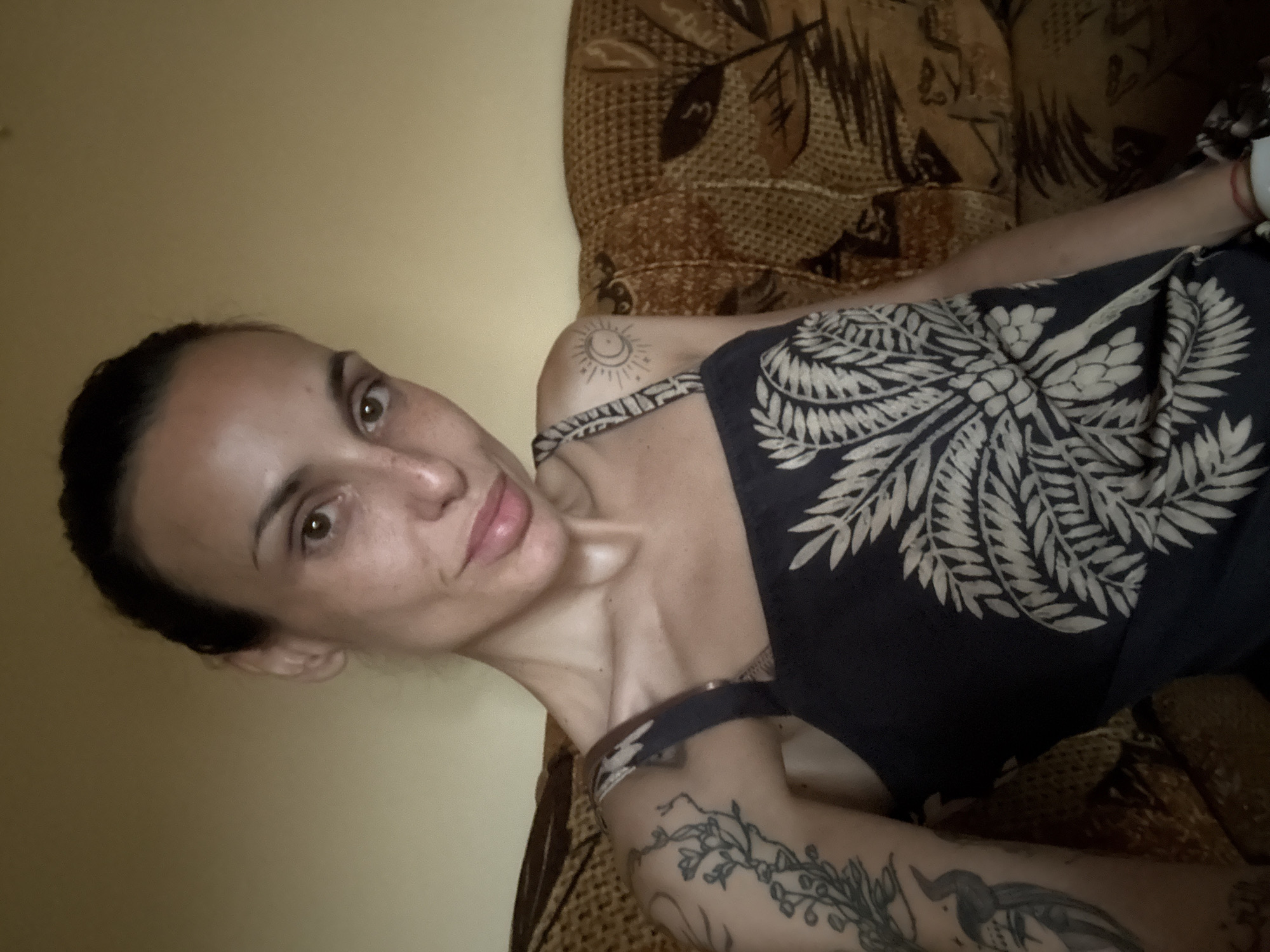 The image, taken in Pleven, Bulgaria, captures a woman lying down indoors, likely on a couch or bed, while looking directly at the camera. Her head is positioned to the left side of the frame, with her body extending diagonally towards the upper right. She has a neutral to subtly pleasant expression, with a slight smile.
The woman has dark hair pulled back. She is wearing a dark-colored top or dress featuring a prominent light-colored pattern of botanical or tribal motifs, resembling leaves or fronds. Several tattoos are visible on her upper body: a stylized sun and crescent moon design on her left shoulder, and more extensive floral or vine-like patterns, alongside another abstract design, on her left arm. A thin, red string bracelet is present on her right wrist.
The setting appears to be an indoor space, possibly a living room or bedroom. Behind the woman, the upholstery of a couch or bed is visible, showcasing a textured brown and beige pattern with leaf-like motifs. The background wall is a plain, light yellowish-brown color. The lighting is soft and ambient, suggesting indoor illumination, but does not indicate a specific time of day. No text is visible in the image.