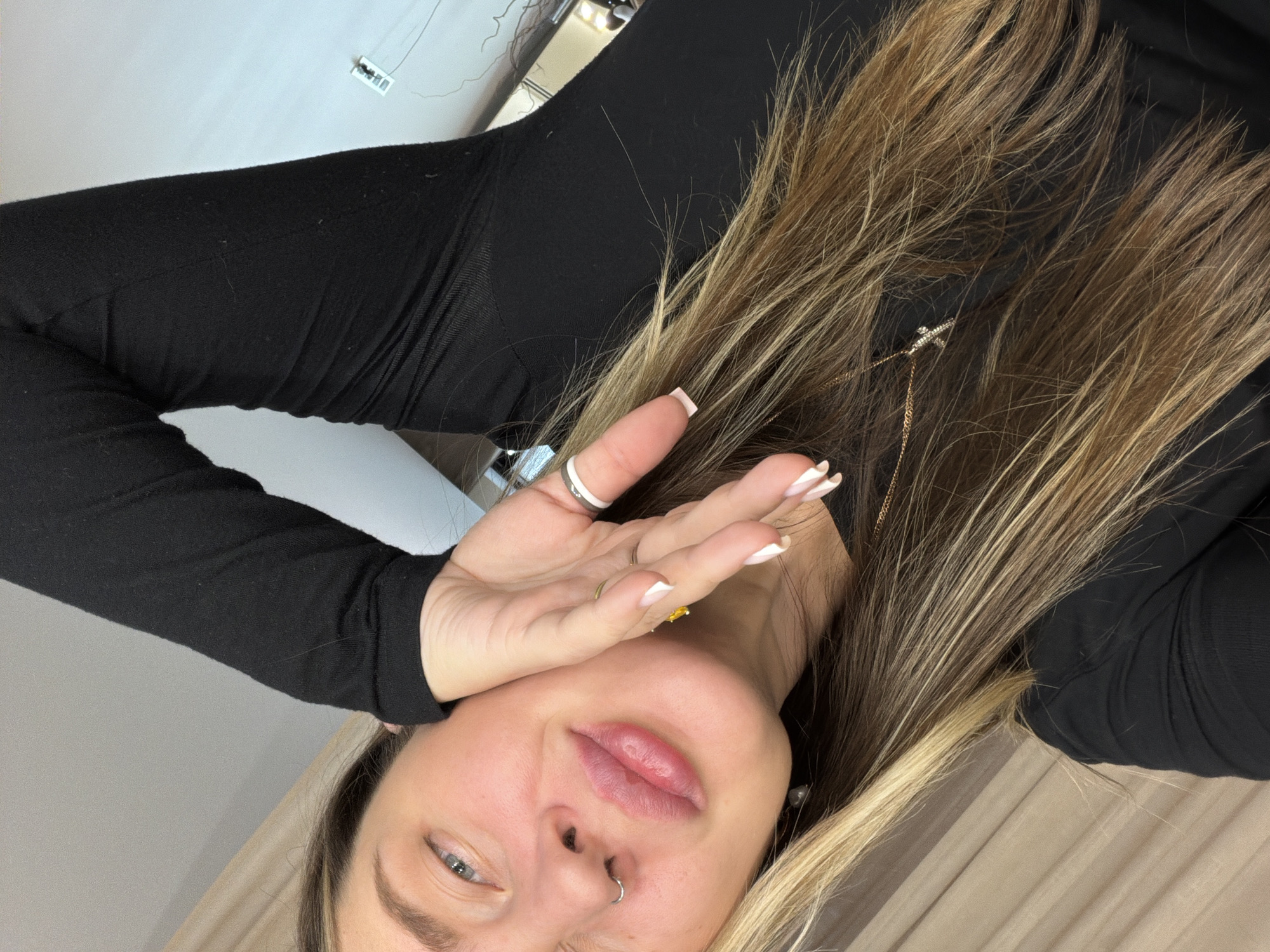 A woman with blonde and brown hair, wearing a black long-sleeved shirt, is lying down indoors. She has fair skin, a nose ring, and is wearing multiple rings on her left hand. Her hand is partially obscuring her face. A gold necklace is visible near her hair. The background includes a wall with electrical wiring and a light fixture. The location is potentially Dolgoprudny, Russia, but there is no visual information that confirms this location.