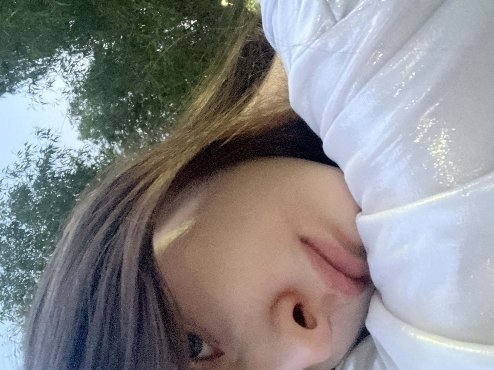 A close-up, diagonal shot features a person, presumably female, resting her head in an outdoor setting in Sinyavino, Russia. Her right eye, nose, and mouth are visible, with her gaze directed slightly upwards. Dark hair frames her face, partially obscuring the forehead. She is wearing a light-colored, possibly white, garment with a subtle, shimmering texture, which she uses as a cushion for her head. The background consists of blurred green foliage from trees against a light blue sky, indicating daylight conditions.