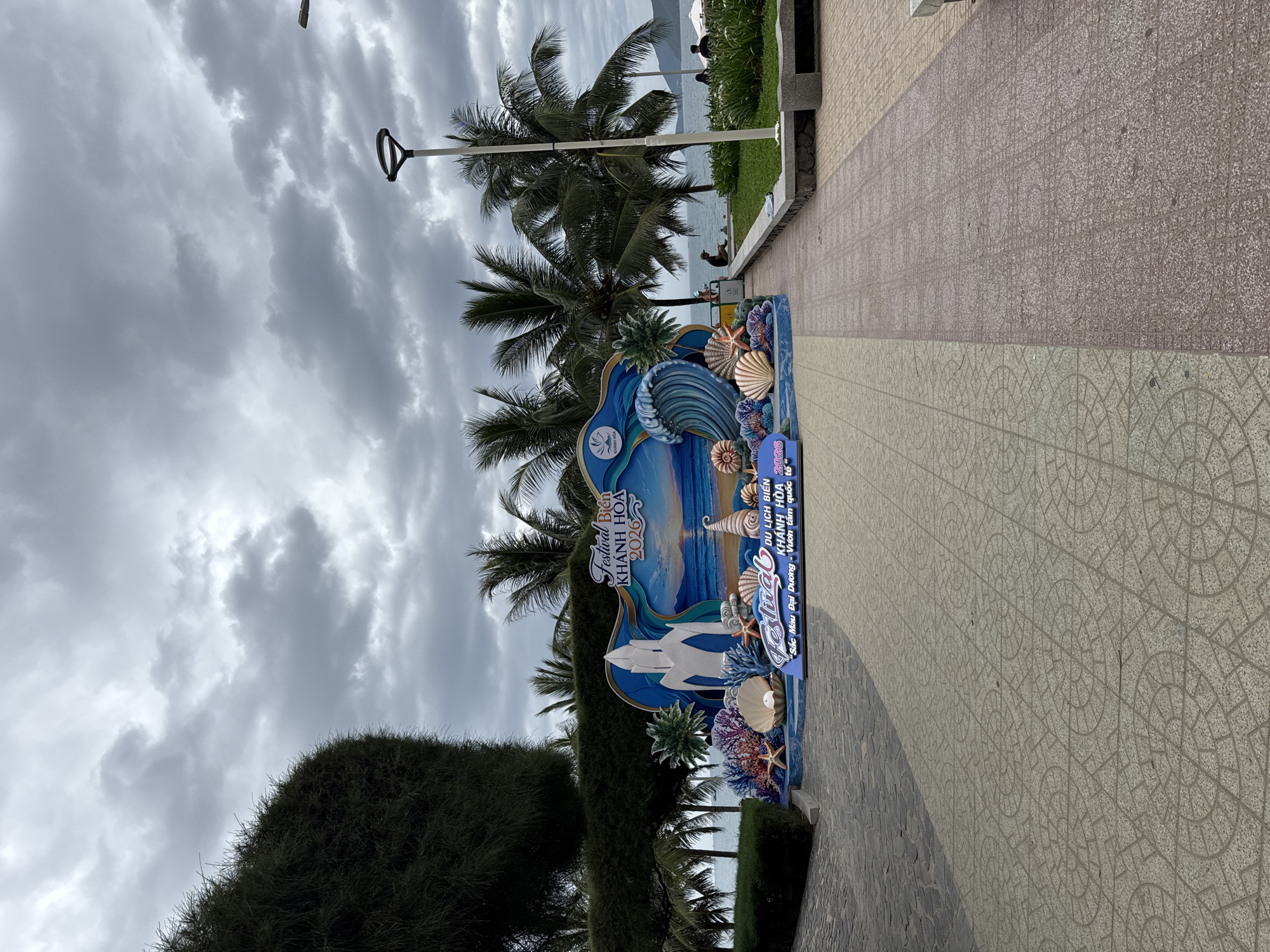 This image depicts a coastal scene in Nha Trang, Vietnam, featuring a prominent, colorful stage setup for a festival. The stage, designed with a wave motif, showcases an abstract white structure, coral, seashells, and starfish. Text on the stage reads "Festival Bien KHANH HOA 2026" and "Sắc Màu Đại Dương - Vườn Tâm Quốc Tế". Palm trees line the background, with the ocean visible beyond. A paved walkway with a patterned surface leads towards the beach where several individuals are seated on benches and the sand. The sky is overcast with dramatic clouds.