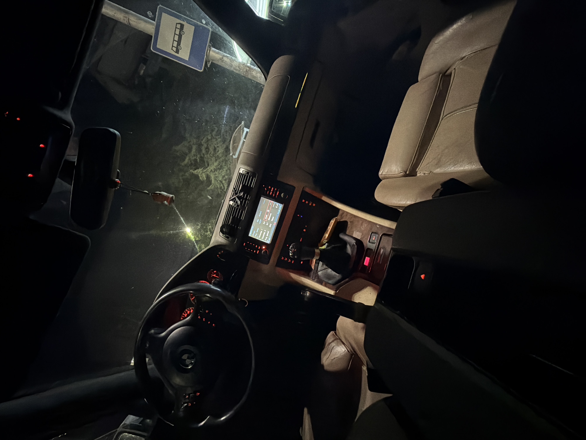 The image captures the interior of a car at night, viewed from the driver's perspective looking forward and slightly to the right. The car's dashboard and central console are illuminated, contrasting with the dark exterior.
Key objects inside the car include a black steering wheel with a visible BMW logo, an illuminated dashboard displaying gauges with red backlighting, and a central digital screen showing vehicle information such as "Driv," "Avg," "Range," "Time," and the date "01/04/2023." Below the screen are various climate control vents and buttons. To the right, a gear shifter is visible. The passenger seat, made of light beige or tan leather, is partially visible on the right side. A rearview mirror hangs in the center, from which a small red and white object dangles. A red indicator light is also visible on the driver's door panel.
Through the windshield, a blue rectangular road sign with a white icon of a bus is clearly visible, likely indicating a bus stop or bus lane. The external environment appears dark, possibly showing reflections on the windshield from moisture or ambient light. What appears to be dark, textured ground or overgrown vegetation is visible outside the car. A faint light source is present in the upper right background.
The car is stationary, and no individuals are visible. The overall ambiance suggests a quiet nighttime scene in Burgas, Bulgaria.