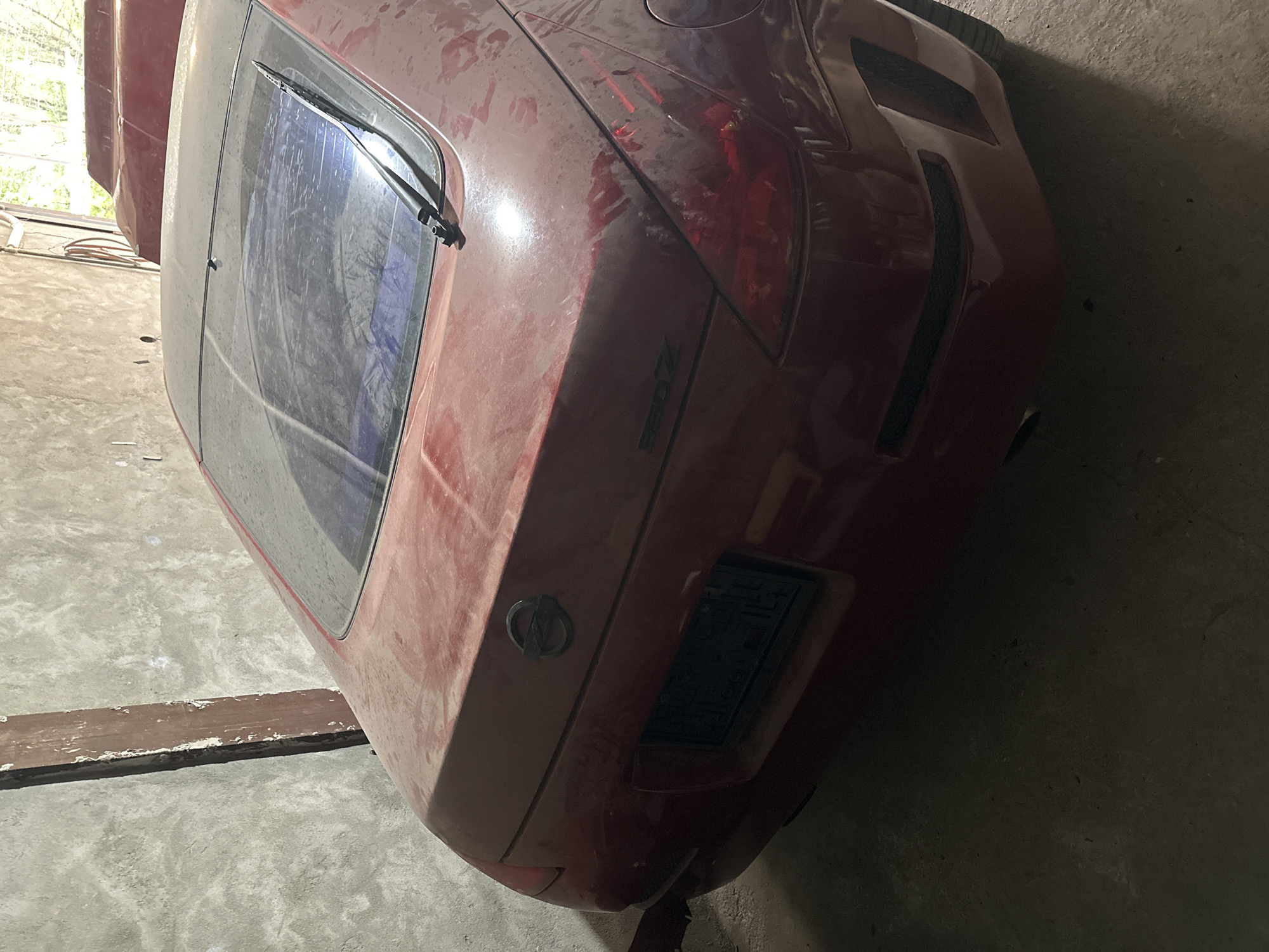 The image shows the rear of a red, possibly maroon-colored, Nissan 350Z coupe. The vehicle appears dusty and is parked indoors, potentially in a garage or storage space. The car's rear window has a wiper, and the Nissan logo is visible on the trunk. The rear taillights are partially visible, as is the "350Z" emblem. There's a license plate holder, but the license plate itself is illegible. The environment appears to be a dimly lit, concrete-floored area with a wall in the background. There's also a wooden plank visible on the left side of the frame. The image lacks any direct evidence to confirm its location is in Yerevan, Armenia. The description is purely based on visible elements in the image.