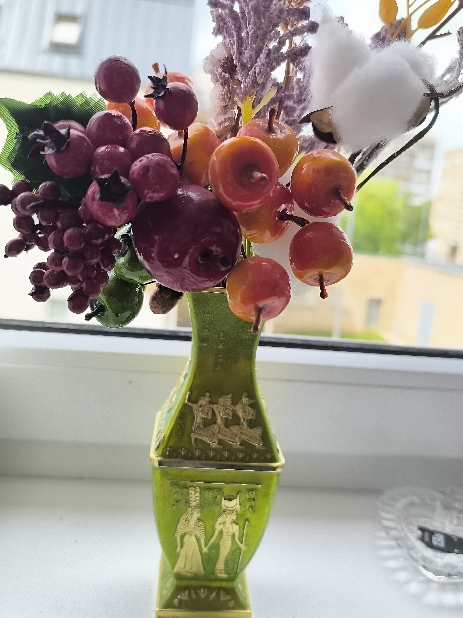 This media file features a close-up view of a decorative arrangement of artificial fruits and natural elements in a green vase, placed on a windowsill. The primary subjects are clusters of red berries, small red and orange apples, and a sprig of cotton. The vase itself is a notable object, adorned with embossed golden figures in an Egyptian style, including depictions of people and hieroglyphs.
The arrangement appears to be a decorative piece, possibly for autumn or seasonal decor. The background shows a blurred view of buildings and trees through a window, suggesting an outdoor scene beyond the immediate setting. The lighting suggests it is daytime, with natural light illuminating the subjects. There are no people visible in the frame, and no specific activity is taking place beyond the static display.
The location cues are general, showing an urban or suburban environment outside the window. The overall mood is still and decorative. No discernible text is visible in the image, apart from the hieroglyphic-style embossments on the vase. The placement on a windowsill implies an indoor setting with a view to the outside.