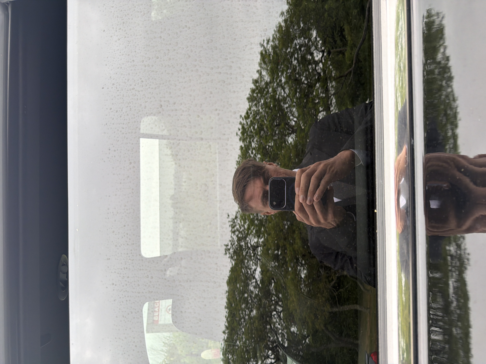 A man in a black suit and tie is reflected in a rain-streaked window, holding a smartphone to take the photo. In the background, a sign reads "BEACH." The scene is outdoors, with trees visible in the reflection, suggesting a location in a park or near a beach in the Town of Southampton, United States of America. A reflection of the lower half of the man's body and the car's exterior is visible on the polished surface below the window.