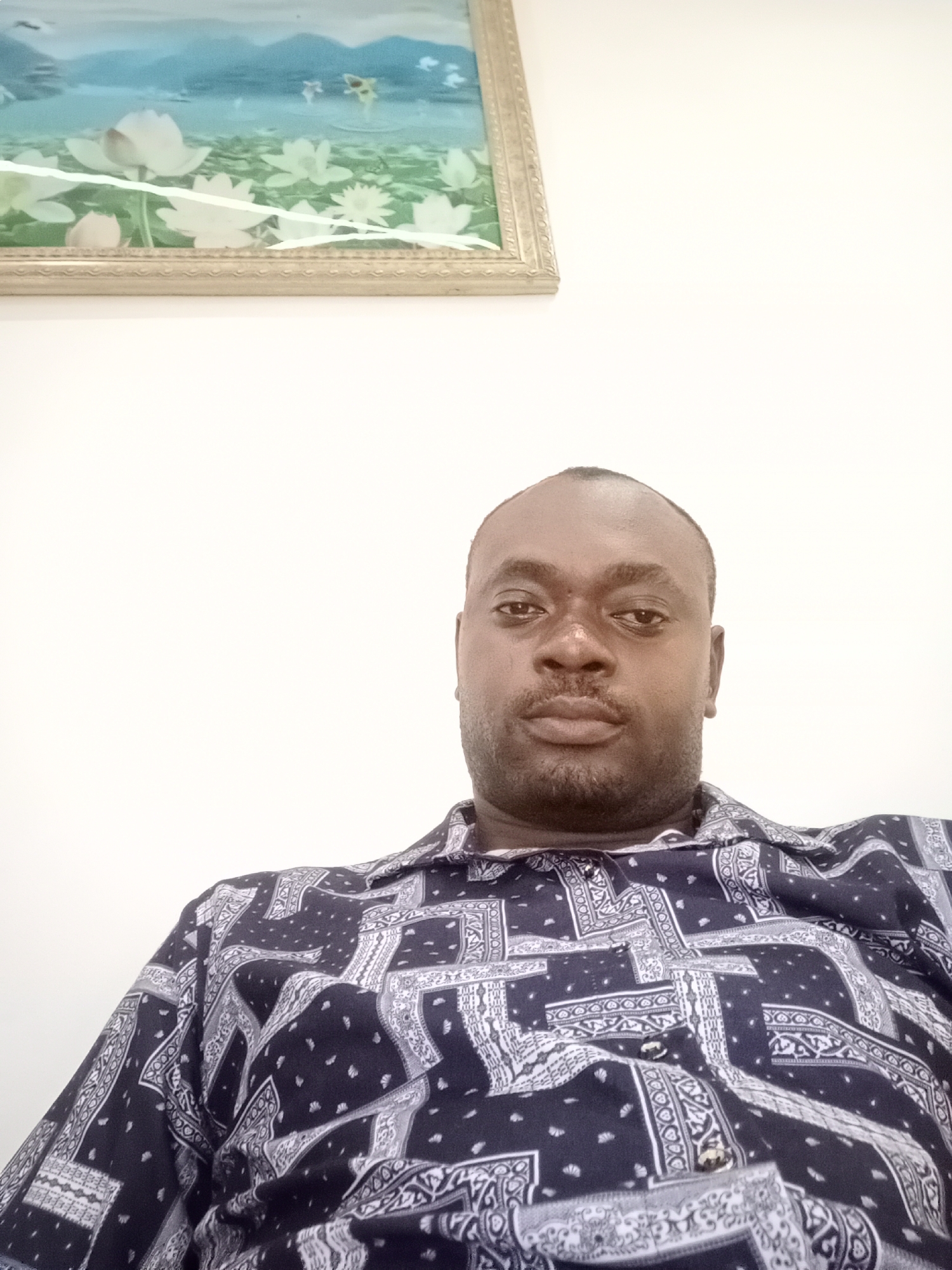 The image shows a medium shot of a dark-skinned man with short hair and a stubbly beard. He is wearing a patterned dark blue and white shirt. Above him on the wall is a framed picture featuring a landscape or water scene. The location is identified as Gwarinpa, Nigeria.