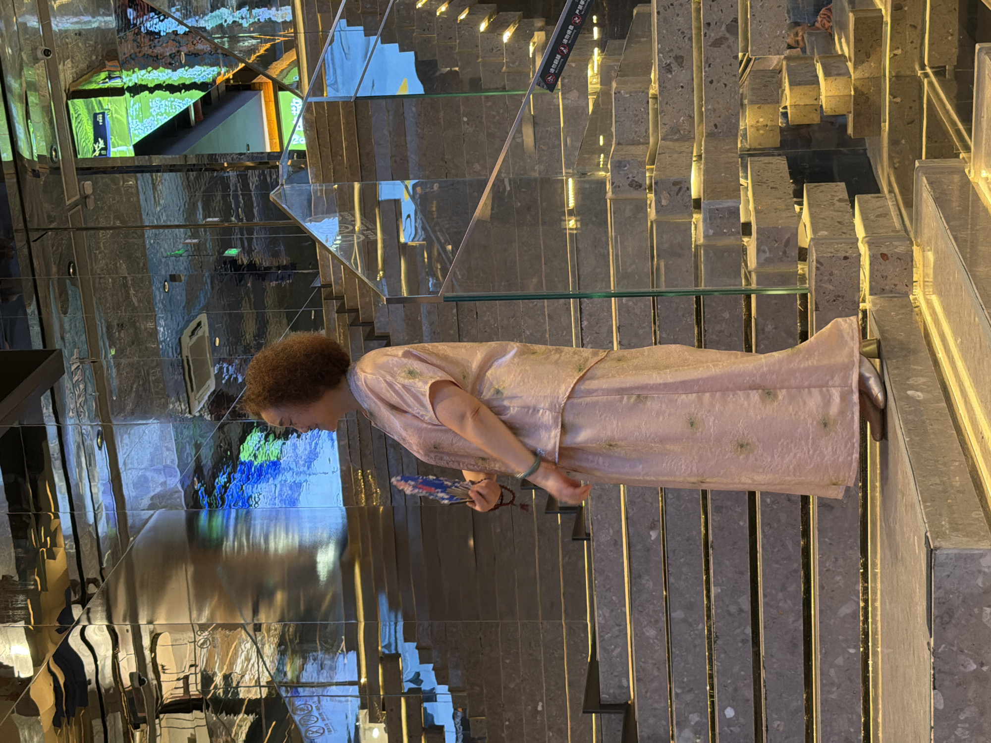 The image shows a woman standing on a wide, modern staircase in what appears to be a public indoor space. The staircase is constructed from large, textured concrete-like blocks with embedded lighting. The woman is wearing a light pink, flowing outfit with a subtle floral pattern and is holding a decorative fan. Her hair is curly and dark, and she is looking towards the right of the frame. The background features a highly reflective, possibly mirrored, surface that distorts and mirrors the staircase and surrounding elements, creating a disorienting and abstract visual effect. This reflective surface is angled and faceted, suggesting a contemporary architectural design. The overall impression is one of a stylish interior with a focus on reflective materials and geometric forms. While the location is stated to be in Yuzhong District, China, this specific detail cannot be visually confirmed from the image itself.
