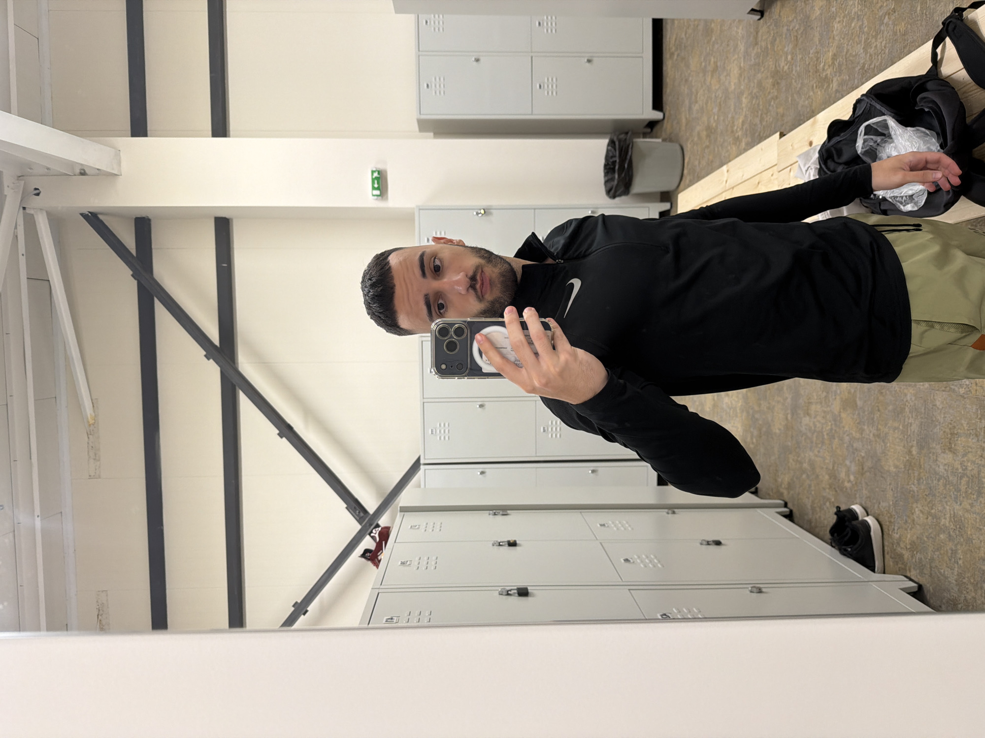 A man is taking a selfie in a locker room. The locker room has gray metal lockers, a trash can, and a wooden bench. A backpack sits on the bench. There is no information to indicate a location in Plovdiv, Bulgaria.