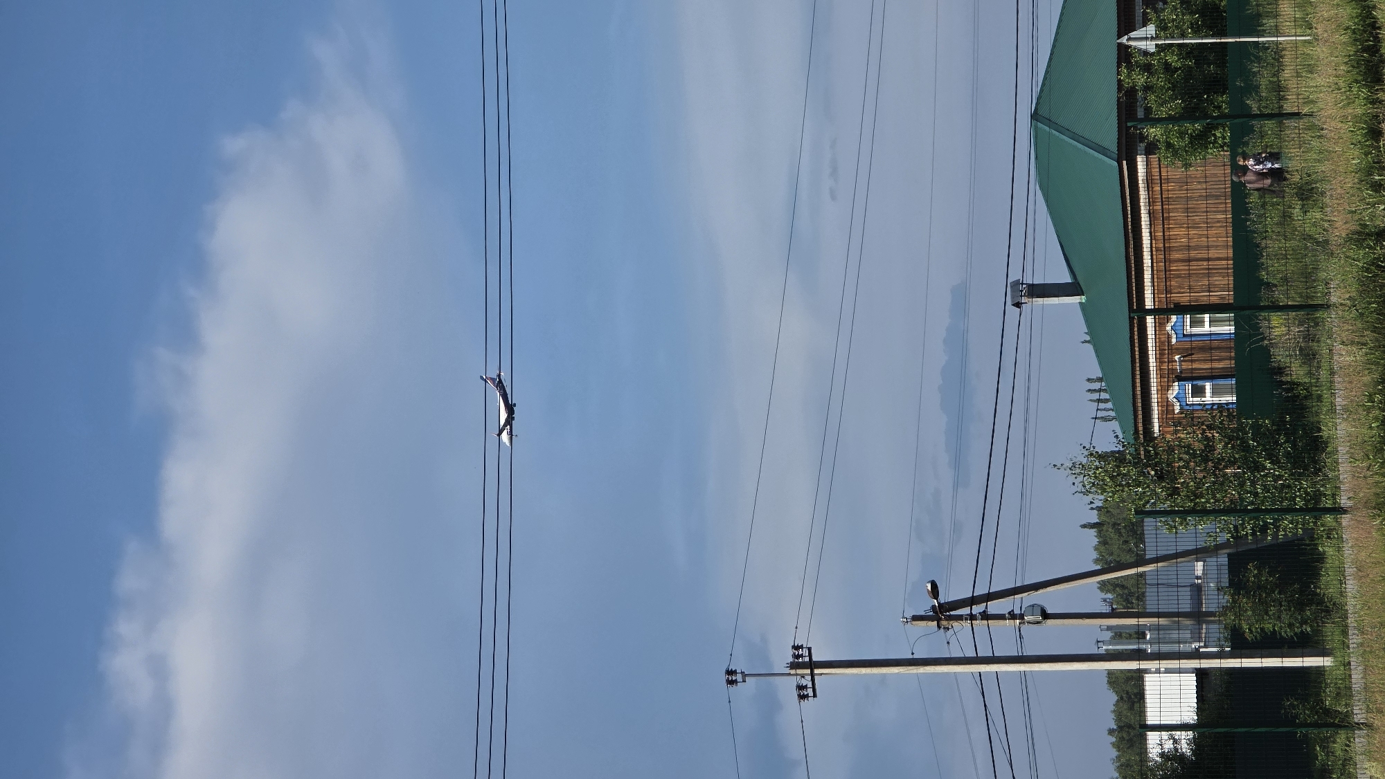An airplane is seen flying in the sky above a residential area with a wooden house, power lines, and a fence in Bulgakovo, Russia. The sky is partly cloudy, with a mixture of blue sky and white clouds. The airplane is a commercial jet with a white fuselage and a blue and red tail. It is flying at a moderate altitude. The house is a single-story wooden structure with a green metal roof. The windows have decorative blue trim. Two people are visible near the house, standing behind a green metal fence. The area is surrounded by trees and grass. Power lines run across the foreground and middle ground of the image, supported by concrete poles.