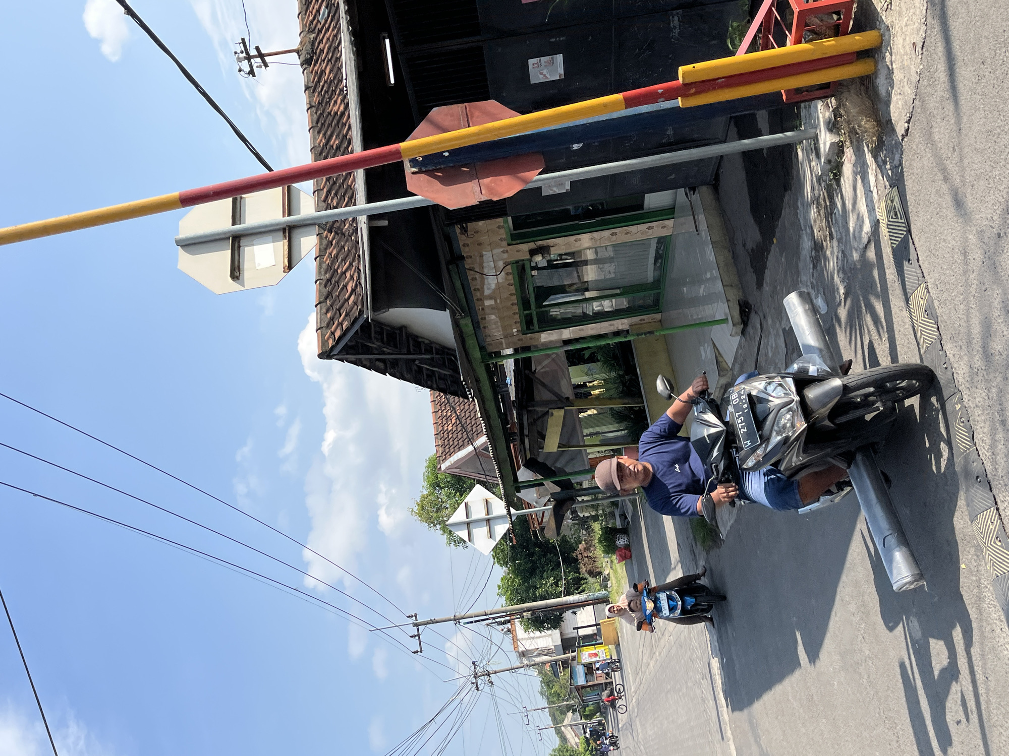 A man wearing a blue t-shirt and shorts rides a black motorcycle down a paved street in Kludan, Indonesia, under a clear blue sky with scattered clouds. The motorcycle has a license plate that reads "W 2757 OB". In front of the motorcycle, there is a speed bump, and the motorcycle is carrying a long, cylindrical object wrapped in silver material. To the left of the man, a woman wearing a headscarf rides a blue motorcycle with a child behind her. In the background, there are several houses with tiled roofs and utility poles with electrical wires. A red and yellow striped pole, possibly part of a railroad crossing, stands on the right side of the frame.