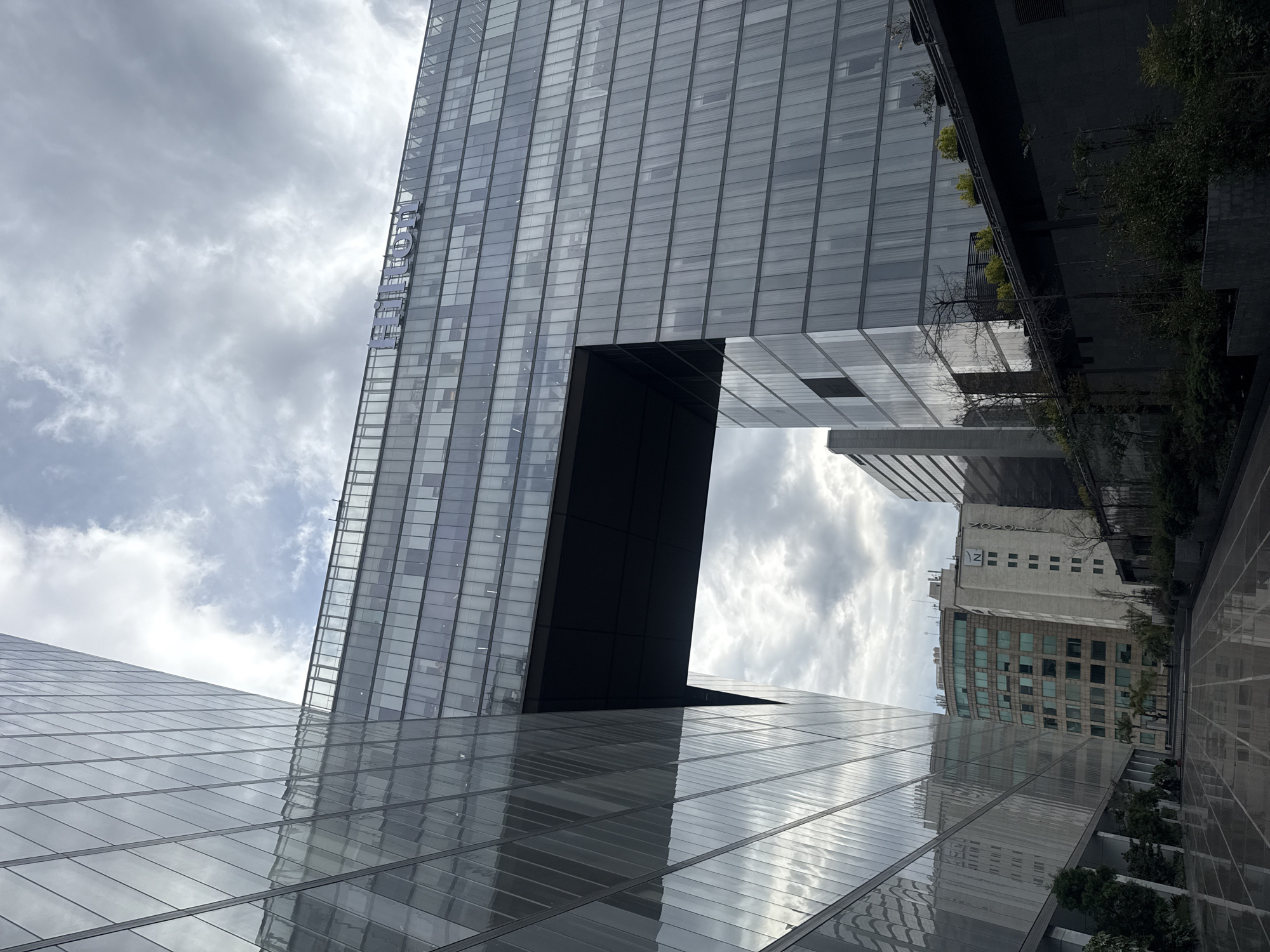 The image shows a view looking upward at modern architecture in Mexico City, Mexico. The primary structure is a tall, glass-facade building with a large void cut out of its midsection. The name "FORUM" is written vertically along the side of the building. The surfaces reflect the cloudy sky. The building is reflective, mirroring other buildings and vegetation within the city. In the background, other buildings are visible. The perspective is from a ground-level position, looking upward at an angle, creating a sense of height and scale.