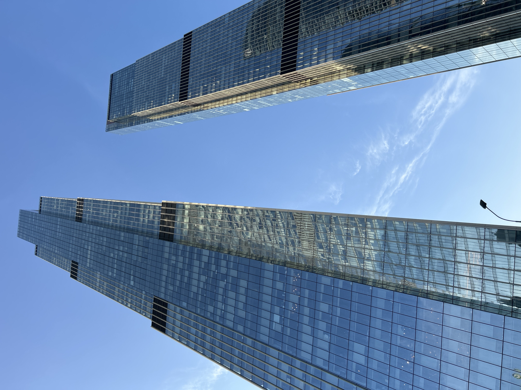 The image is a low-angle shot of two modern glass skyscrapers against a clear blue sky. The buildings appear to be located in a large city. One skyscraper is positioned at the top of the frame, while the other is located towards the bottom. The buildings have many windows reflecting the sky and possible neighboring buildings. A street light is visible next to the bottom skyscraper.