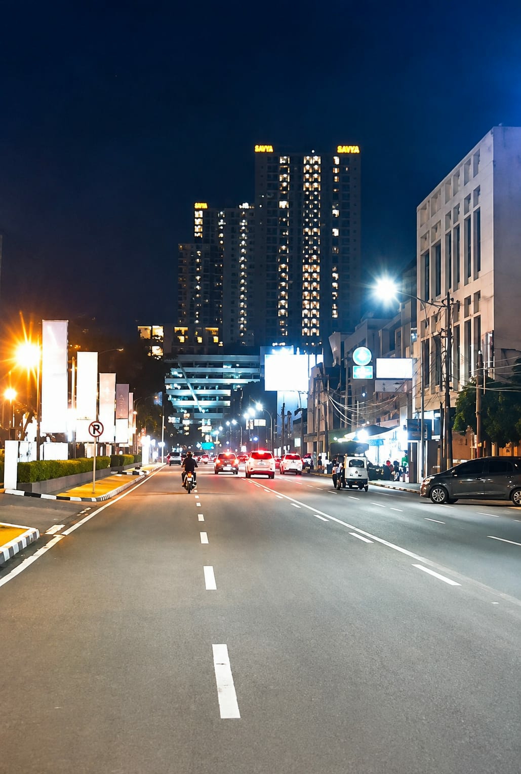 This is a nighttime, low-angle, exterior, medium shot of a wide city street in Medan, Indonesia. The street is well-lit by streetlights and the illuminated windows of buildings. Several vehicles, including cars, a motorcycle, and a three-wheeled auto-rickshaw, are driving on the road. Buildings flank the street, with a prominent high-rise apartment building in the background that is lit up. There are also streetlights, traffic signs, and decorative banners visible. The sky is dark blue, indicating it is nighttime.