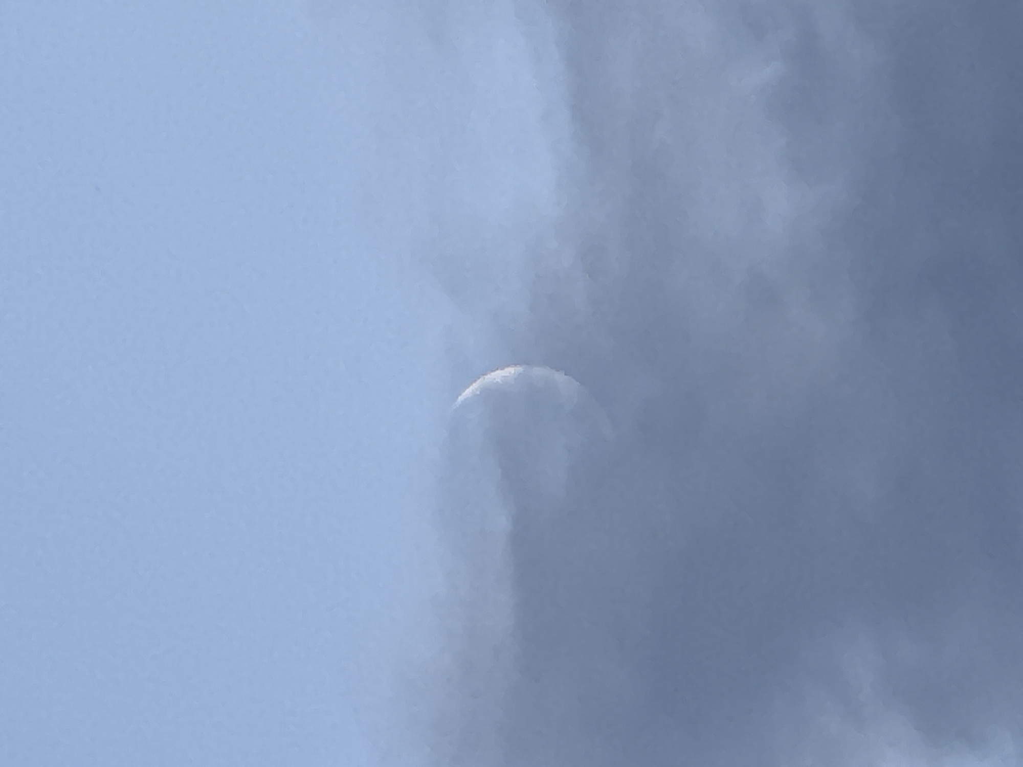A crescent moon is visible through a layer of dark clouds in a pale blue sky. The moon appears as a thin sliver of light, partially obscured by the cloud cover. The clouds are dense and cover a significant portion of the sky, creating a moody and atmospheric scene. The image was taken in the city of Chaikovsky, Russia.