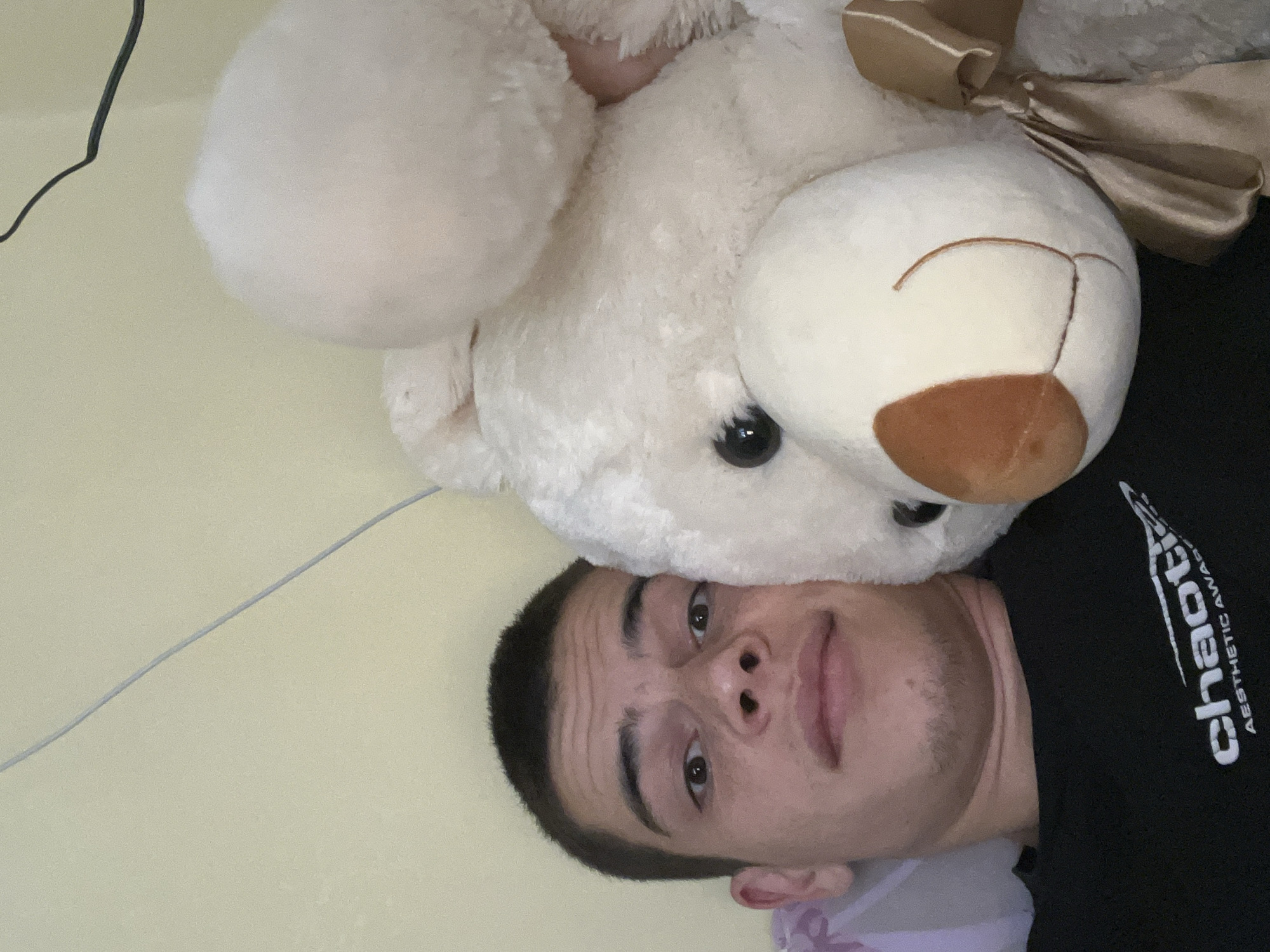 A selfie-style photograph features a light-skinned individual with dark hair, their face positioned alongside a large, white teddy bear. The bear, which appears to be lying on top of the person, has a brown nose and a satin bow tied around its neck. The individual is wearing a black t-shirt with the word "chaotic" printed in white, along with smaller text that reads "AESTHETIC AWARE". The background is a plain, light-yellow wall with a power cable partially visible at the top left. The photograph is taken at an angle, rotating the scene. Without any specific landmarks or identifying details, it's not possible to determine if the photo was taken in Plovdiv, Bulgaria or anywhere else for that matter.