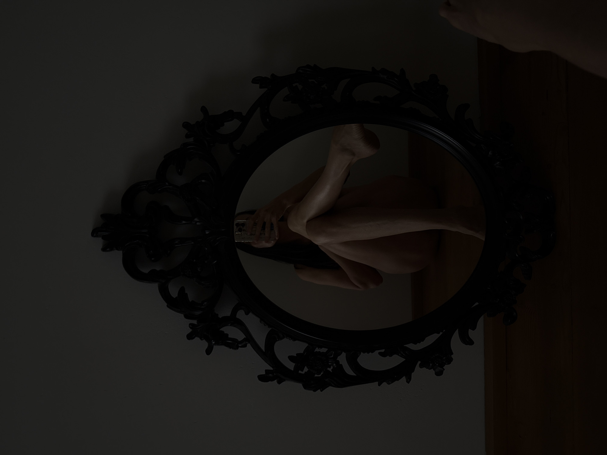 The image depicts a person's legs and torso reflected in an ornate, black oval mirror. The person is positioned such that their body is contorted into a fetal-like pose, with knees drawn up and arms wrapped around them. One foot is clearly visible in the reflection, with its sole facing upwards. The lighting is dim, suggesting an indoor setting possibly at night or in a dimly lit room. The mirror's frame is intricately designed with floral and scroll motifs, adding a vintage or gothic aesthetic to the scene. The background is dark, with a hint of a wall on the left and a wooden surface on the right, potentially a door or furniture. A hand, possibly belonging to the person in the reflection, is visible at the top right corner of the original image, partially obscuring a lighter area. The overall mood is introspective and perhaps melancholic, amplified by the low lighting and the solitary reflection. There is no visible text in the image. The location context of Chaikovsky, Russia, does not provide any specific visual cues within the image itself.