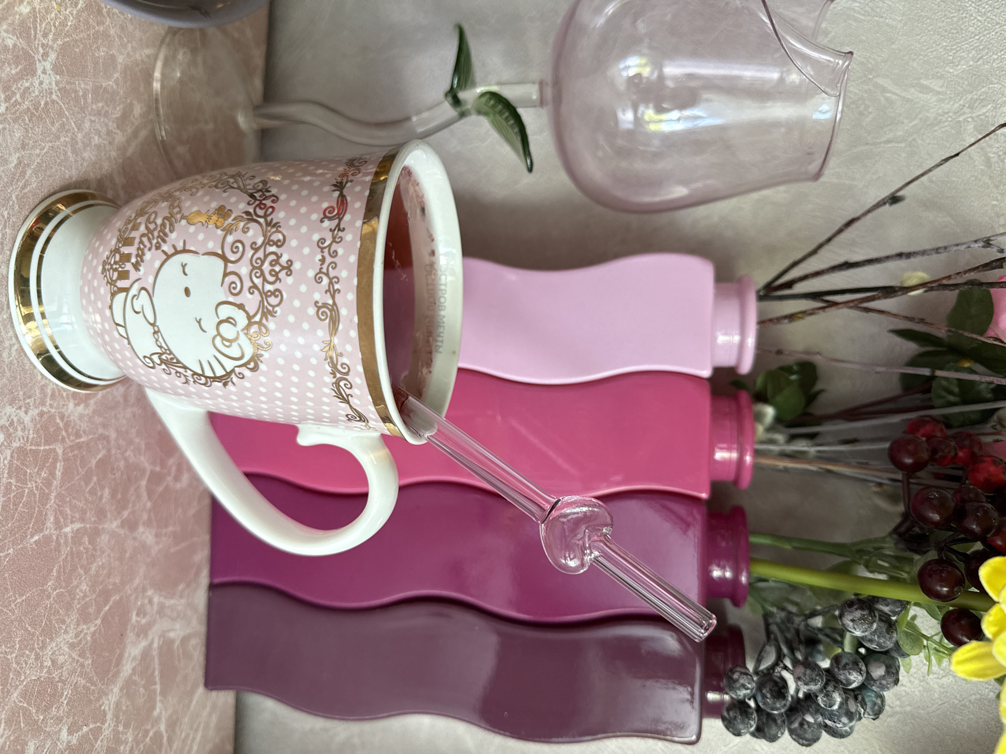 A Hello Kitty mug filled with a pink beverage stands on a pink, marble-patterned surface. A clear glass straw with a heart-shaped accent is inserted into the mug. Behind the mug, a row of wavy vases in varying shades of pink and purple holds decorative branches and artificial berries. To the left of the mug, a delicate pink glass vase with a green leaf accent is positioned. The background is a textured, light-colored wall. The scene does not provide any information that would allow for the determination of a location in the city of Chaikovsky, Russia.