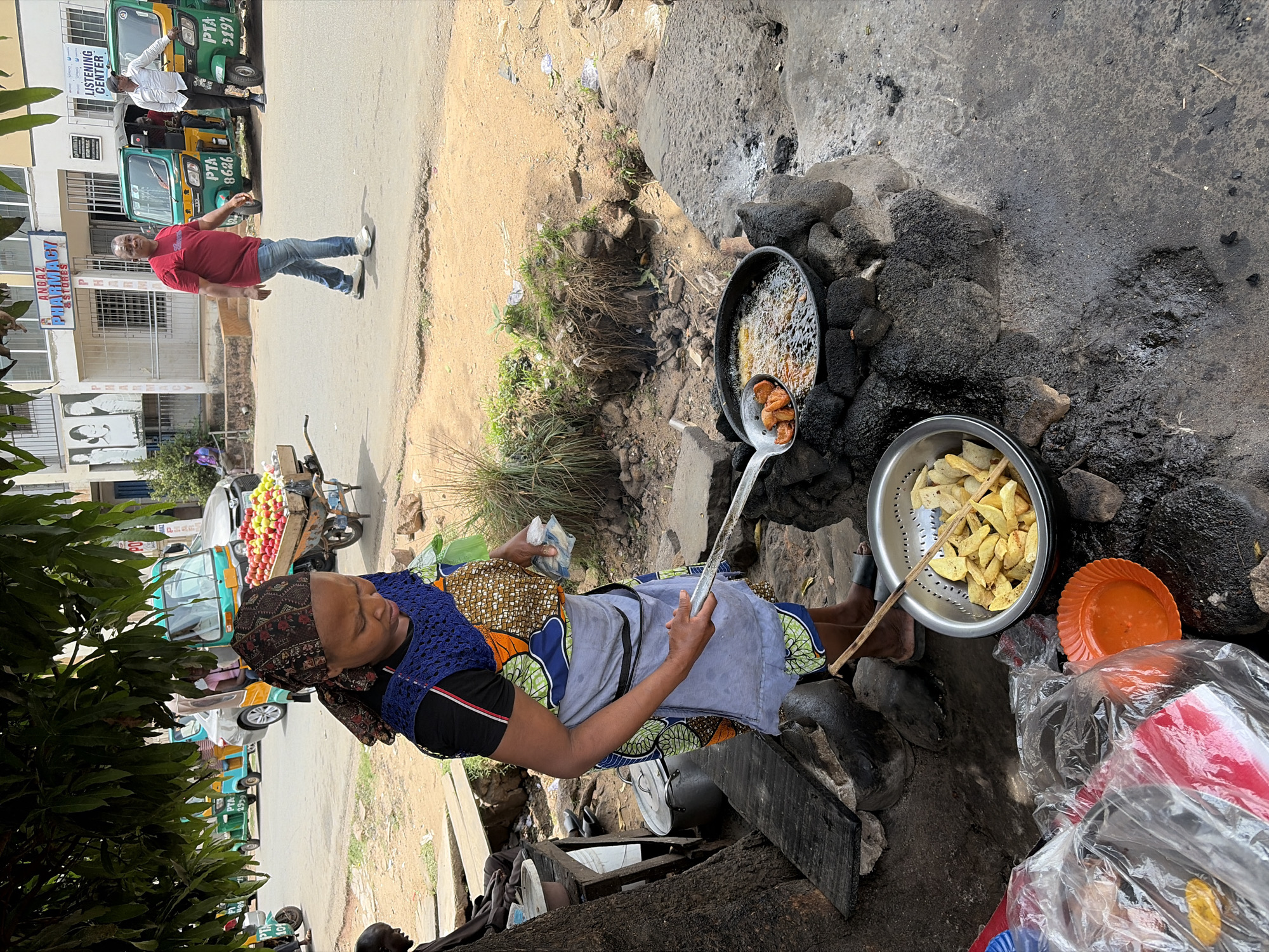 An outdoor street scene in Jos, Nigeria, depicts a woman cooking street food alongside a busy road during daytime. In the foreground, a woman wearing a patterned headscarf, a blue crocheted vest over a black t-shirt, and a colorful apron/skirt is actively frying food. She holds a long-handled skimmer, tending to food (likely yam or potato chips) bubbling in a large black pan set over an open fire constructed with dark rocks and charcoal. Next to the pan, a metal colander holds freshly fried, golden-brown food. An orange plastic plate and clear plastic bags with various items are also visible nearby.
On the paved road behind her, a man in a red t-shirt and blue jeans walks past. Further down the street, green and yellow tricycles (known as keke NAPEPs) are parked or in motion. One tricycle prominently displays "PTA 3391" and another "PTA 9626." A person is visible standing by one of these tricycles. In the background, a building features a sign that reads "ANDAZ PHARMACY & STORES," and another sign below it indicates "LISTENING CENTER." A fruit vendor's cart laden with red and green apples is also seen on the street. The weather appears sunny with clear skies. The overall setting is a vibrant urban environment with improvised street vending and daily public transport.