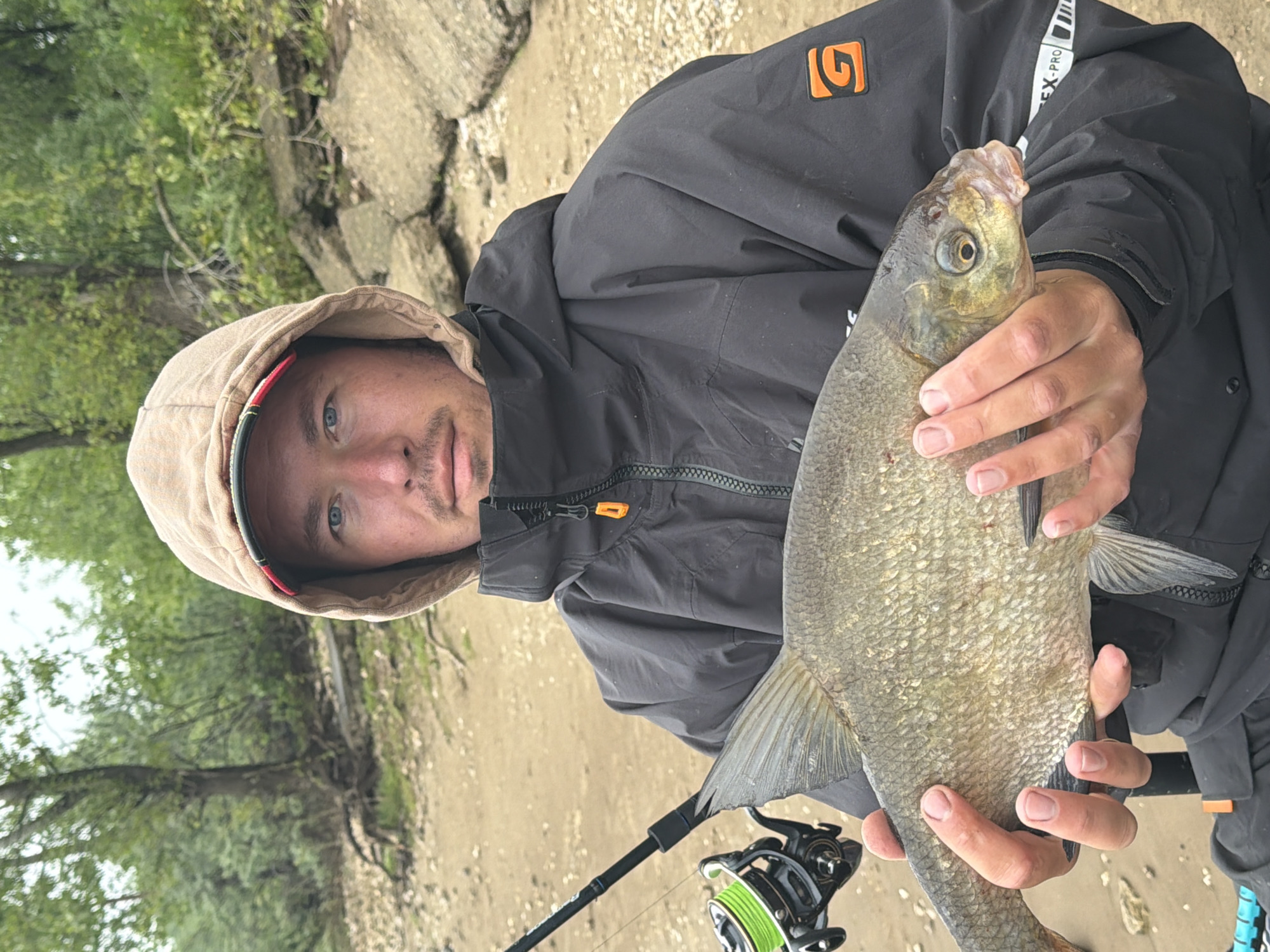 An outdoor photograph, likely taken on a river or lake bank in Noviy, Russia, features a man holding a large fish. The man, viewed from the chest up, has light blue eyes and light facial hair. He is wearing a dark, possibly waterproof jacket with the hood pulled up, revealing a hint of red lining. The jacket features an orange square logo with a stylized 'G' on the left chest area and white "REX PRO" text on a black strap near his right shoulder. He holds a substantial, scaly freshwater fish, possibly a bream, with both hands, presenting it towards the viewer. The fish has a silvery-golden hue and its mouth is slightly agape. In the lower left foreground, a black fishing rod and a reel spooled with bright green line are visible. The background consists of a sandy or muddy bank leading to green trees and foliage, suggesting a natural, riparian environment. The diffused light indicates an overcast or cloudy day. The man's expression is neutral to mildly content.