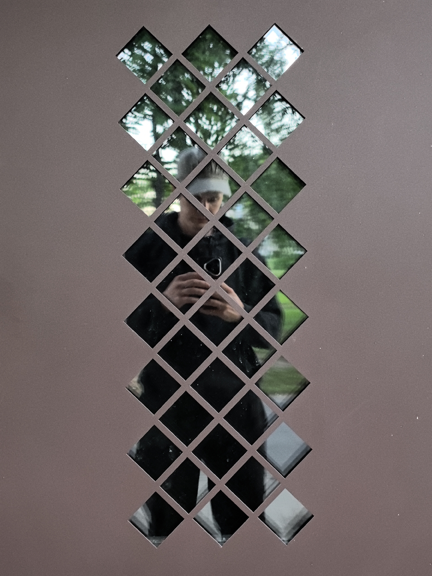 The image captures a reflection of a person holding a phone, looking down at the screen. The person is wearing a dark jacket and a white band on their forehead. The reflection is viewed through a series of diamond-shaped openings in a textured, brown surface. The background visible through the openings consists of greenery and trees, suggesting an outdoor environment. The image appears to be a self-portrait taken through a patterned screen or gate.