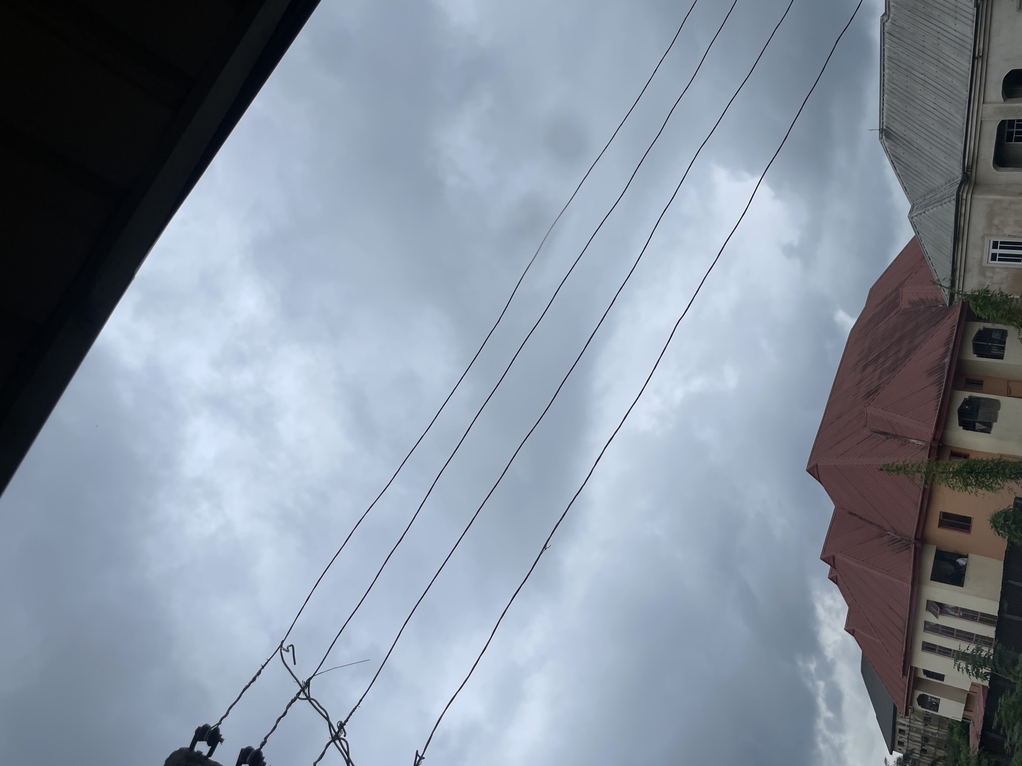 In Ihiagwa, Nigeria, a cloudy sky looms over a residential area. Power lines stretch across the frame, casting dark shadows against the overcast heavens. Below, multi-story buildings with distinct architectural styles are visible. The building in the foreground features a prominent red, corrugated metal roof with multiple gables. Its facade is a blend of light cream and orange, with numerous windows, some open, revealing glimpses of interior spaces. Adjacent to this, a building with a grey, corrugated metal roof is partially visible, showing a lighter-colored facade with window openings. Vegetation, including trees, is present in the lower portions of the scene. The overall atmosphere suggests an overcast day in a densely populated urban or suburban setting.
