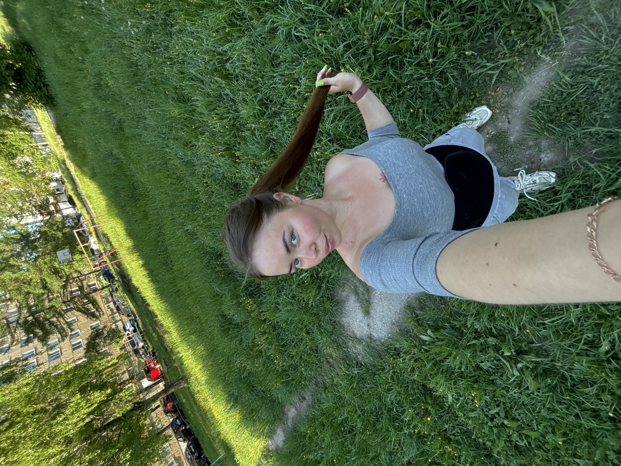 A woman lies on her back in a grassy area in Izhevsk, Russia. She has long brown hair pulled into a ponytail and is holding the ponytail in her right hand, displaying nails painted a bright green color. She wears a gray long-sleeved shirt and gray pants. On the ground next to her are light-colored sneakers. A gold bracelet is visible on her right wrist. In the background, there is an apartment building and a playground. A line of cars is parked along the edge of the grass. The image is taken from a high angle, likely as a selfie.