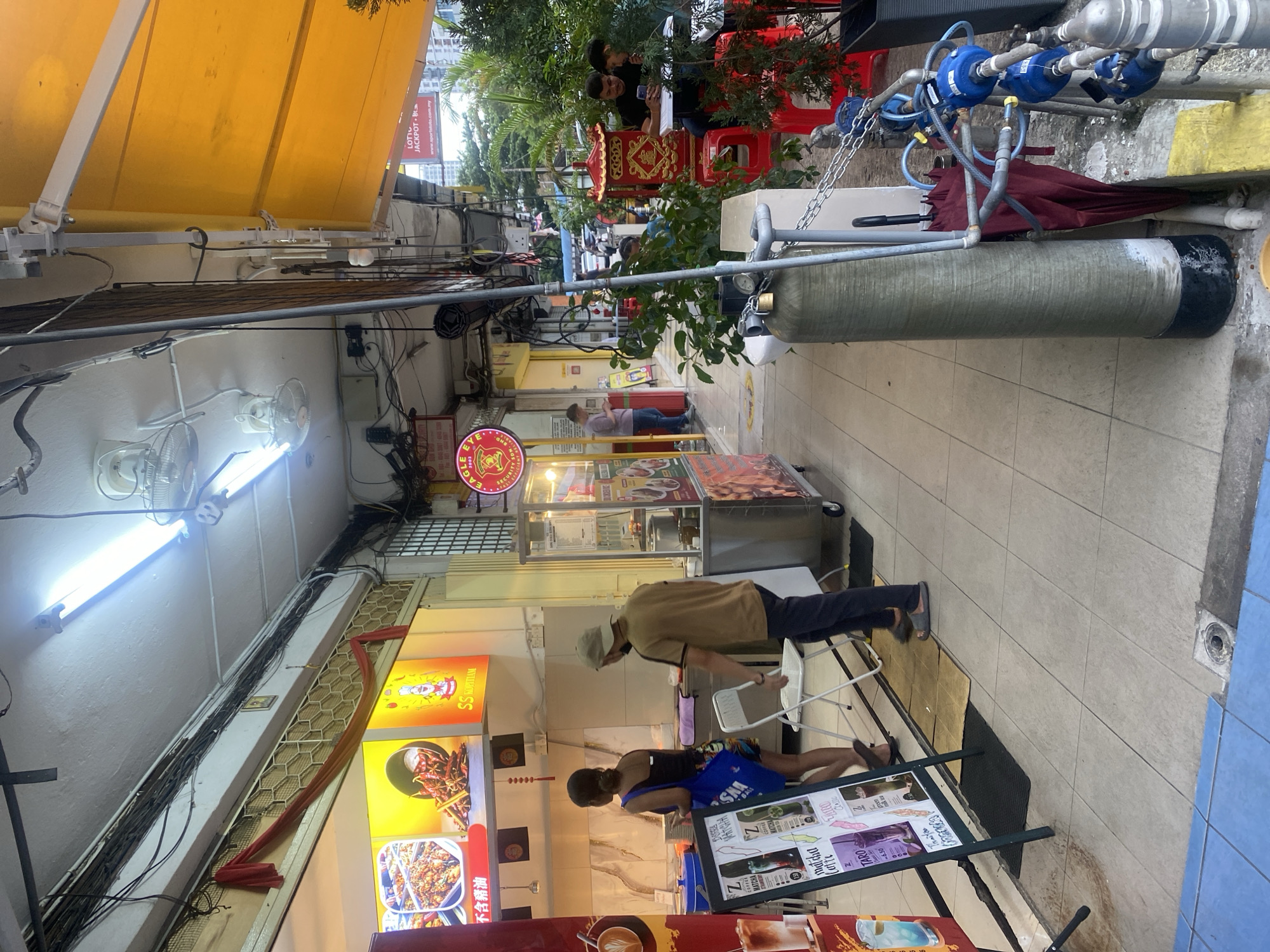 A street-level view captures the exterior of a row of shops in Kuala Lumpur, Malaysia, under a bright yellow awning. In the foreground, a sign displays drink options like matcha and taro. Two women stand near a stall, one with her back to the camera, holding a blue bag. A man in a beige shirt walks away from the viewer. Further down the street, two men are seated at a table outside a shop, looking at a phone, while another person is partially visible behind them. A red umbrella is propped up next to a large cylindrical tank. The scene is illuminated by fluorescent lights and appears to be a bustling commercial area.